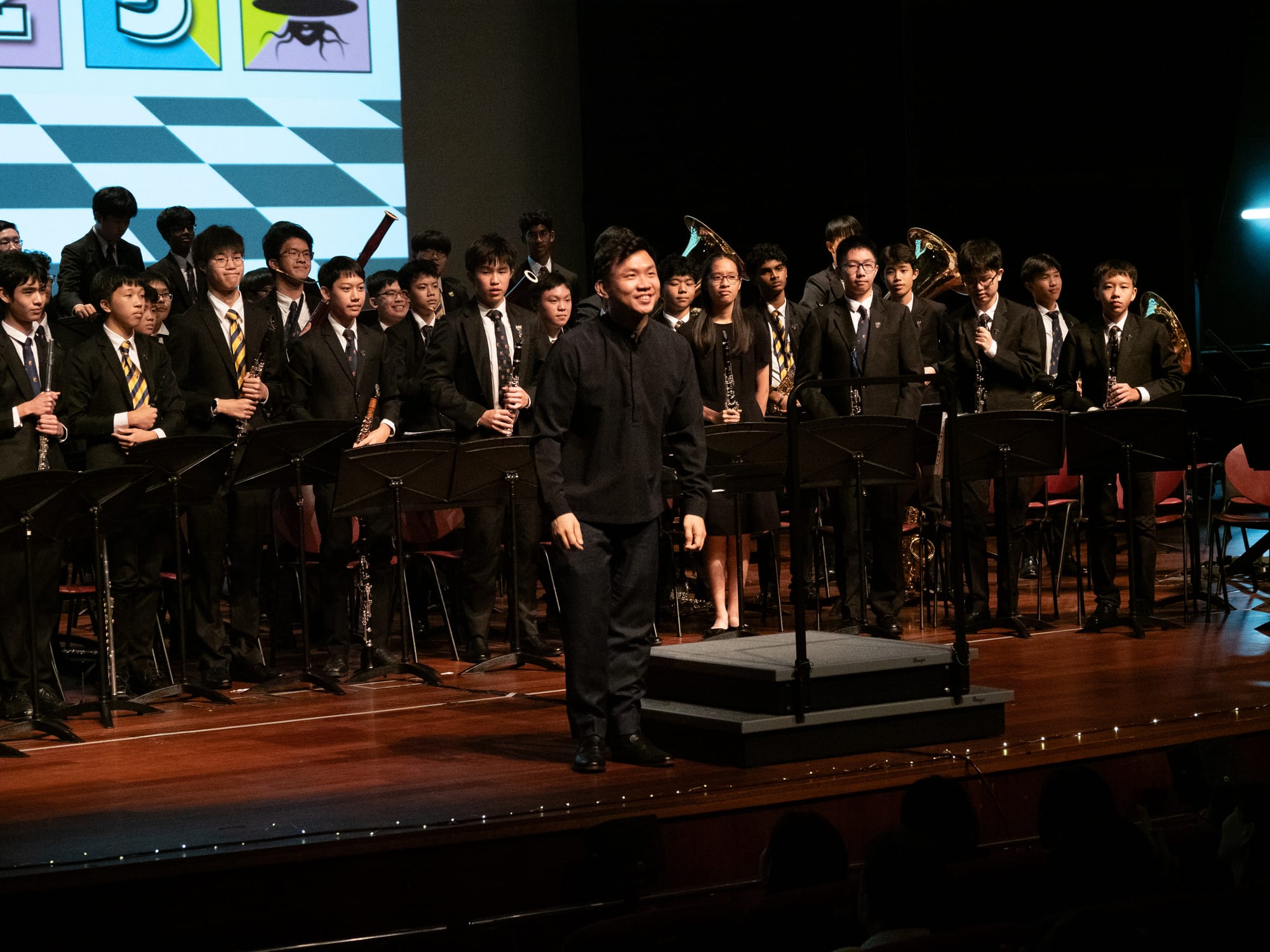 A conductor in black stands before a large youth wind band in formal attire on a concert stage.