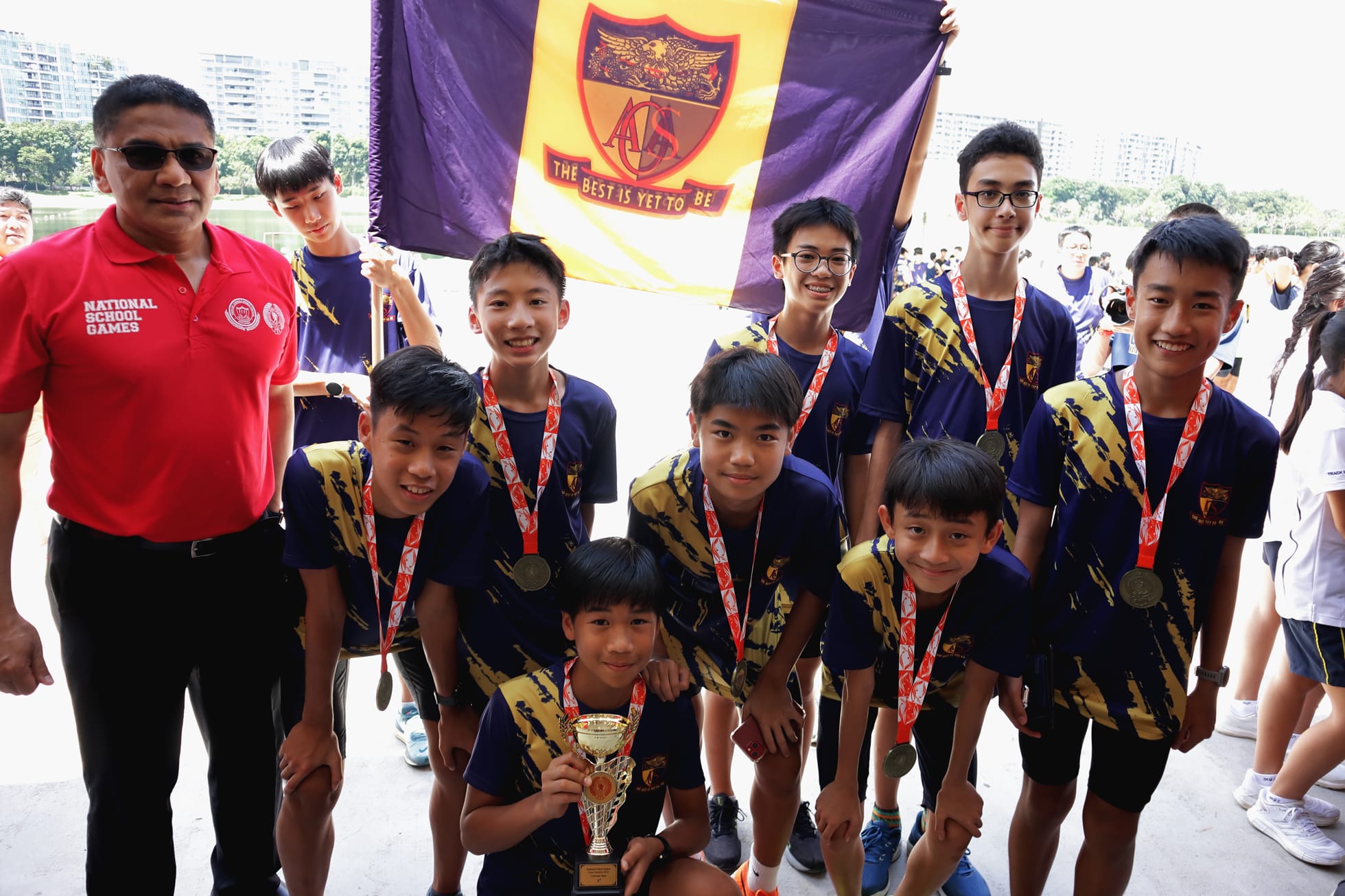 Young students in navy and gold uniforms hold medals and a trophy under a purple ACS school flag at National School Games.