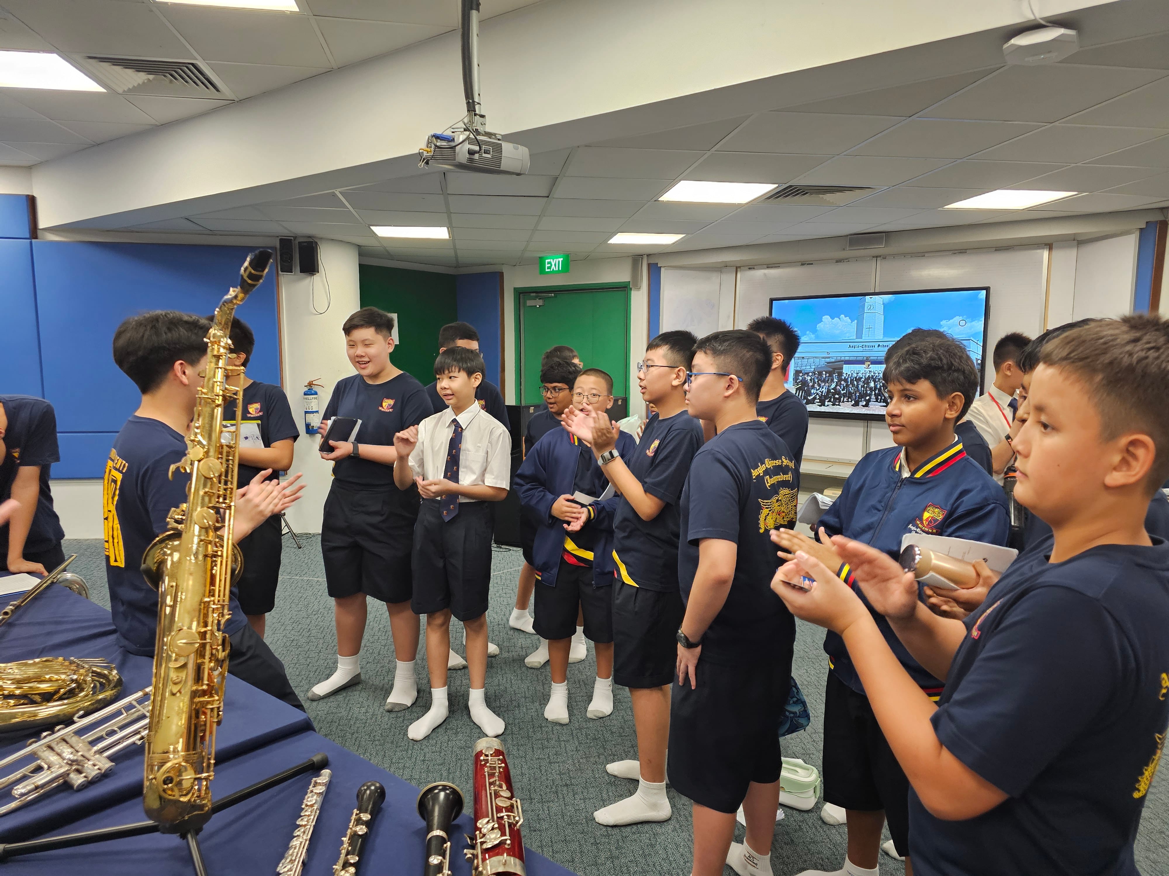 Students in navy uniforms stand in a classroom around a table displaying brass and woodwind instruments including a saxophone.