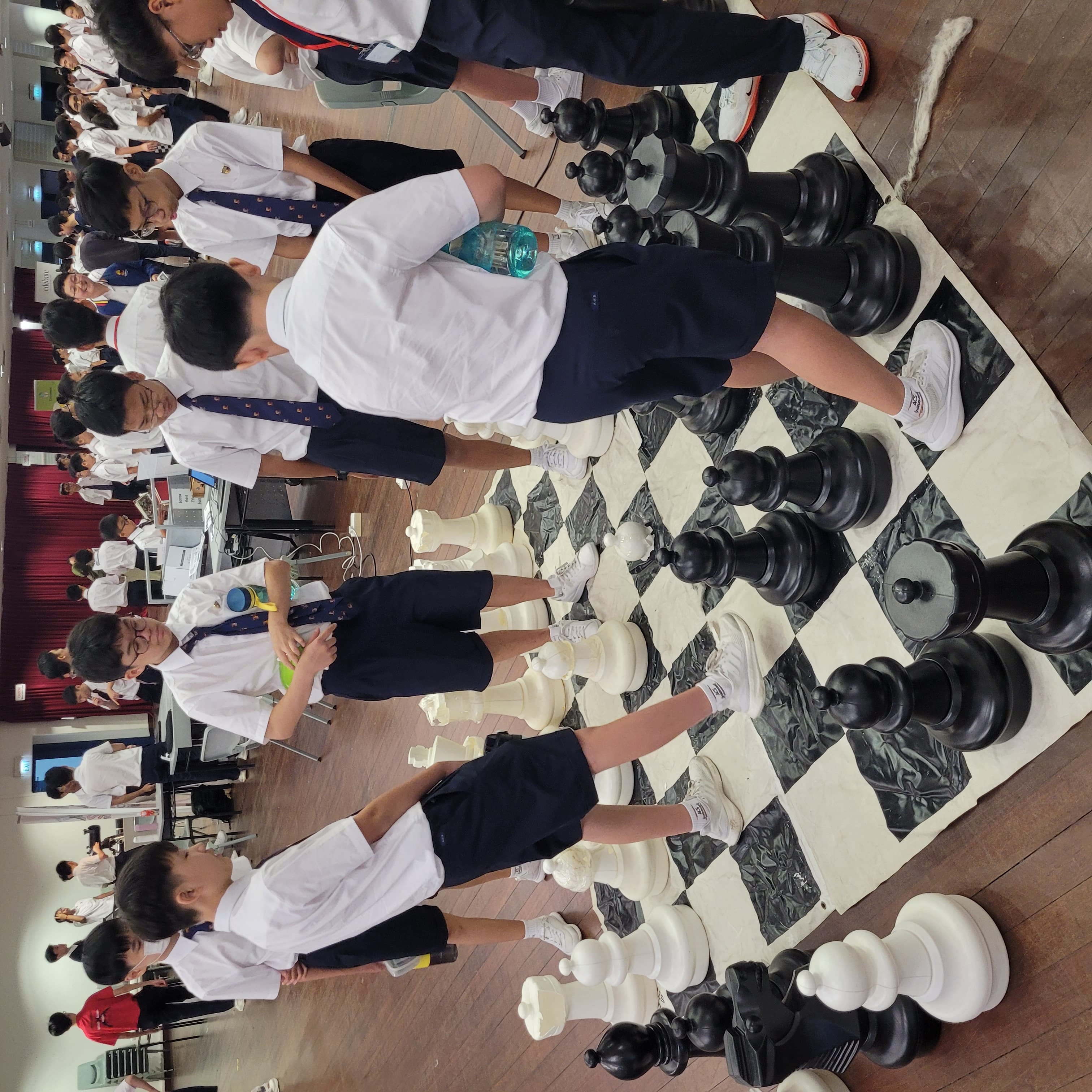 School boys in white uniforms playing on a giant floor chess board with large black and white pieces indoors.