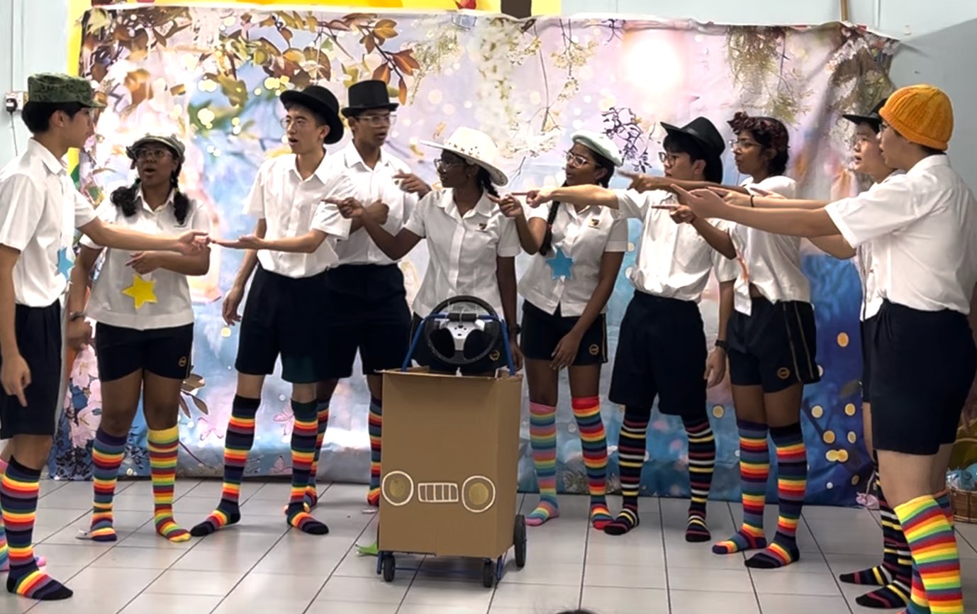 Students in white shirts, black shorts, colorful striped socks, and hats perform a skit around a cardboard car prop.