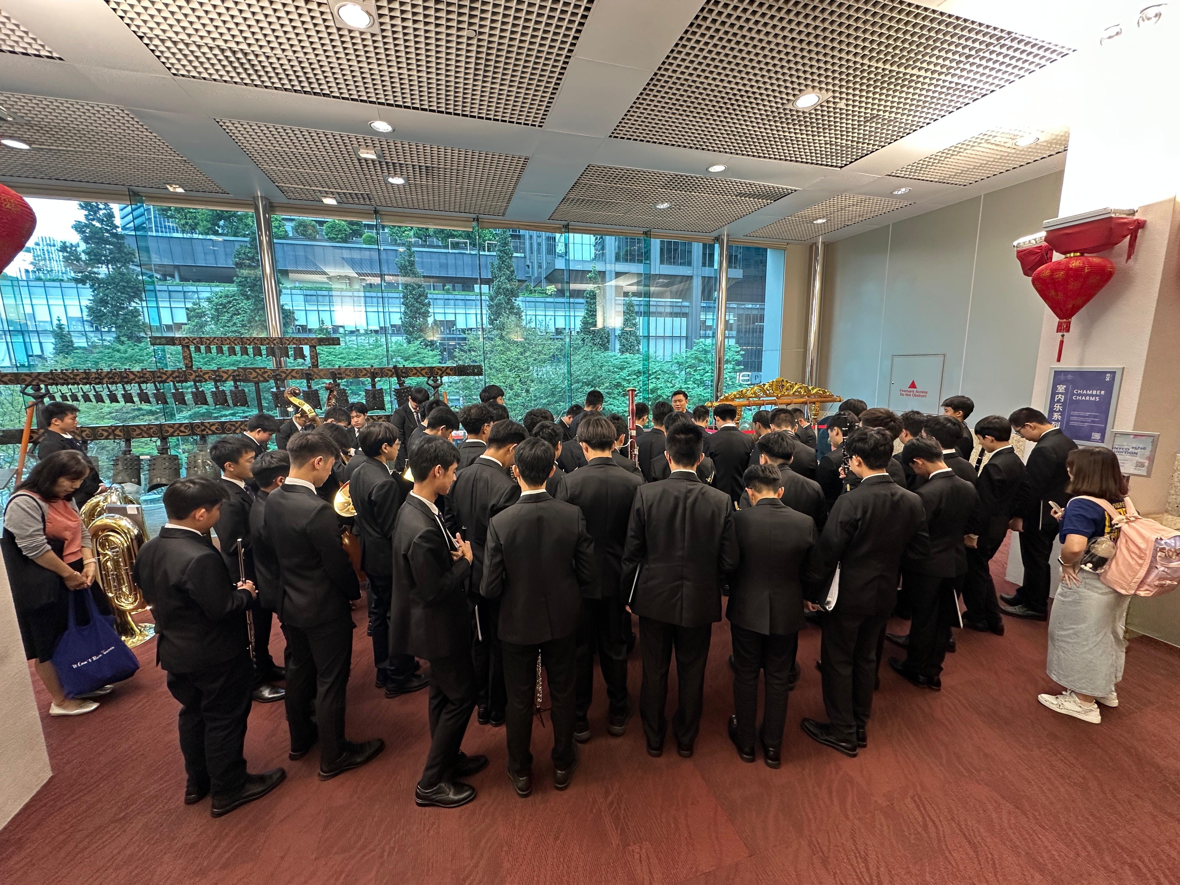 A large group of young musicians in black suits gather indoors near traditional instruments and red lantern decorations.