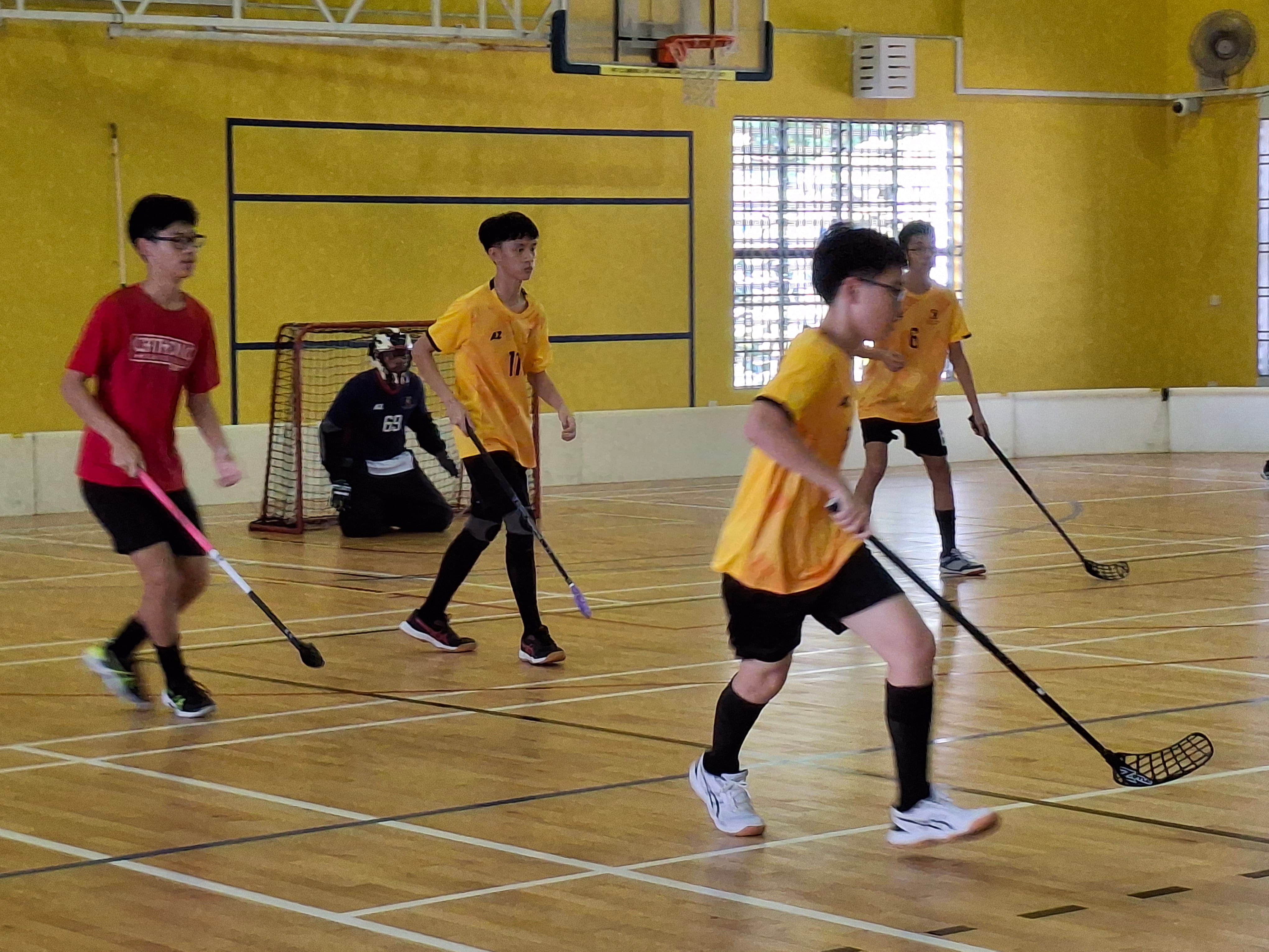 Young players in yellow and red jerseys play floorball indoors, holding sticks near a goal with a goalkeeper.