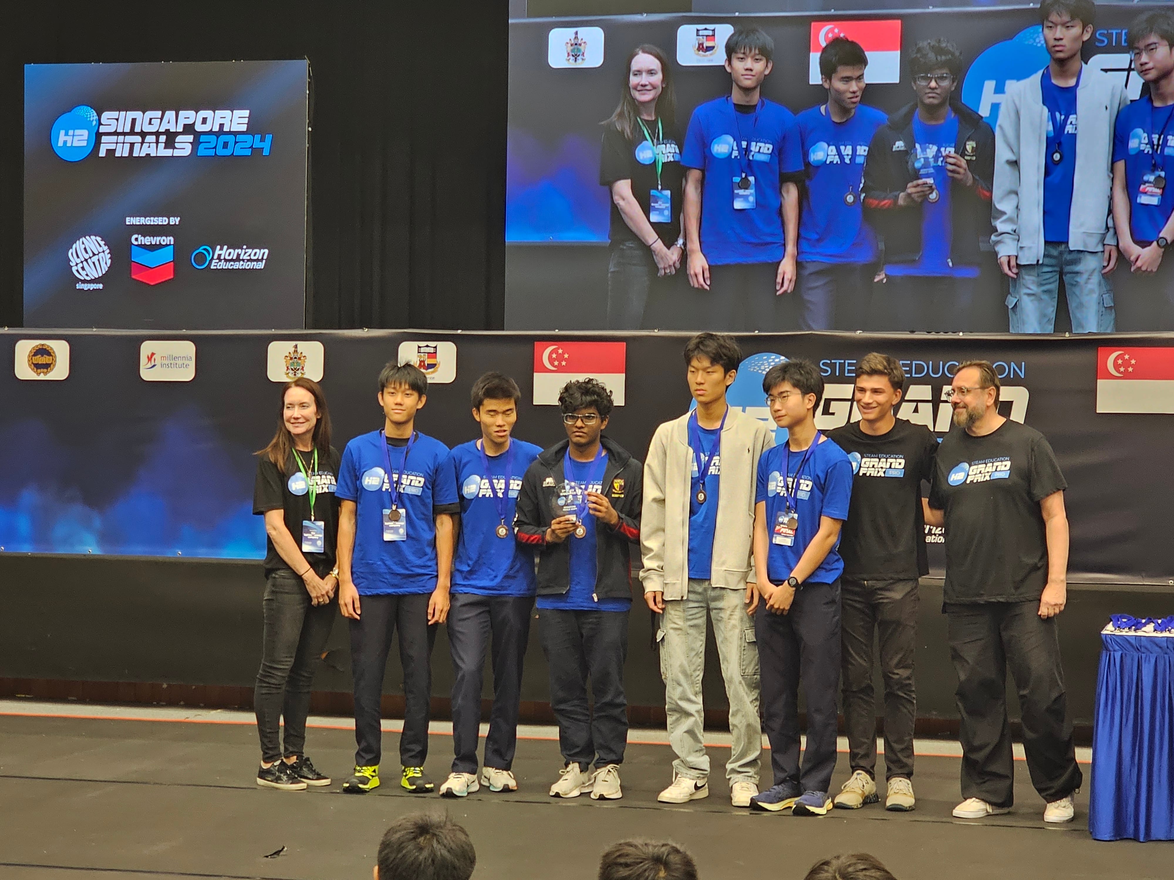 A group of people stand on a stage at the "H2 Singapore Finals 2024". Five of them wear matching blue shirts and lanyards, three of them wear matching black shirts - all of them are smiling proudly. A large screen behind them displays their image. Flags of Singapore are visible, highlighting the event's location. The mood is celebratory and formal.