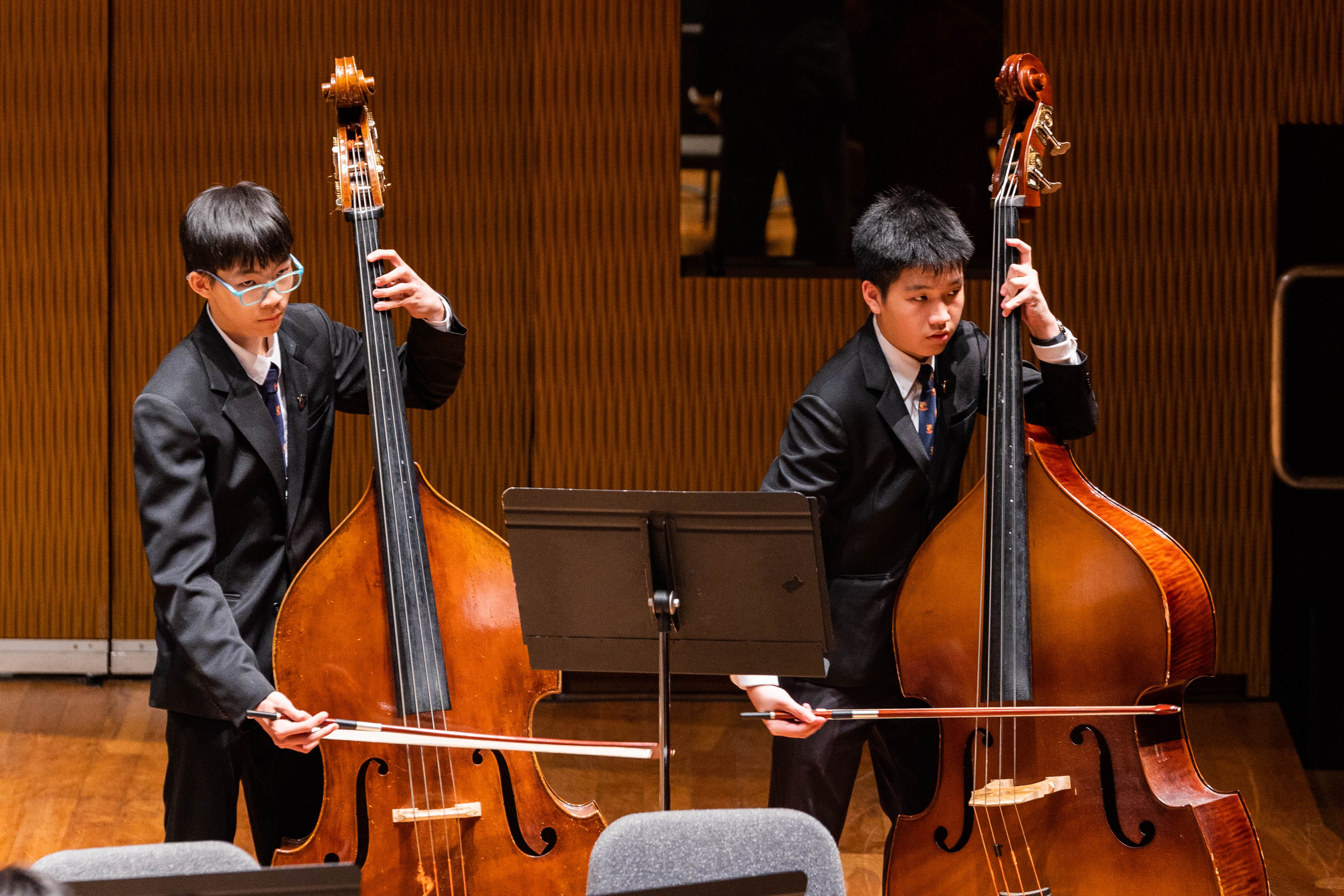 Two young male students in dark suits playing double basses on a wooden stage with a music stand between them.