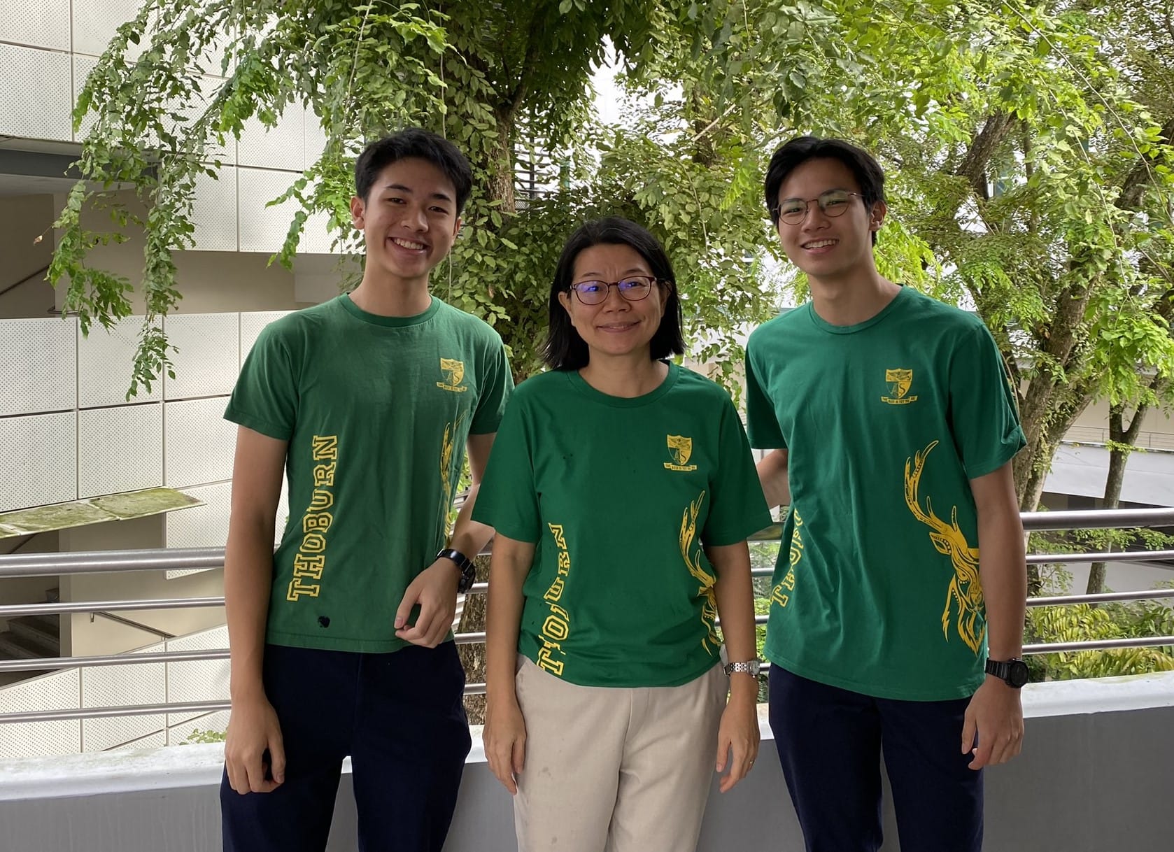 Three smiling people wearing matching green Thoburn T-shirts stand together outdoors near trees and a railing.