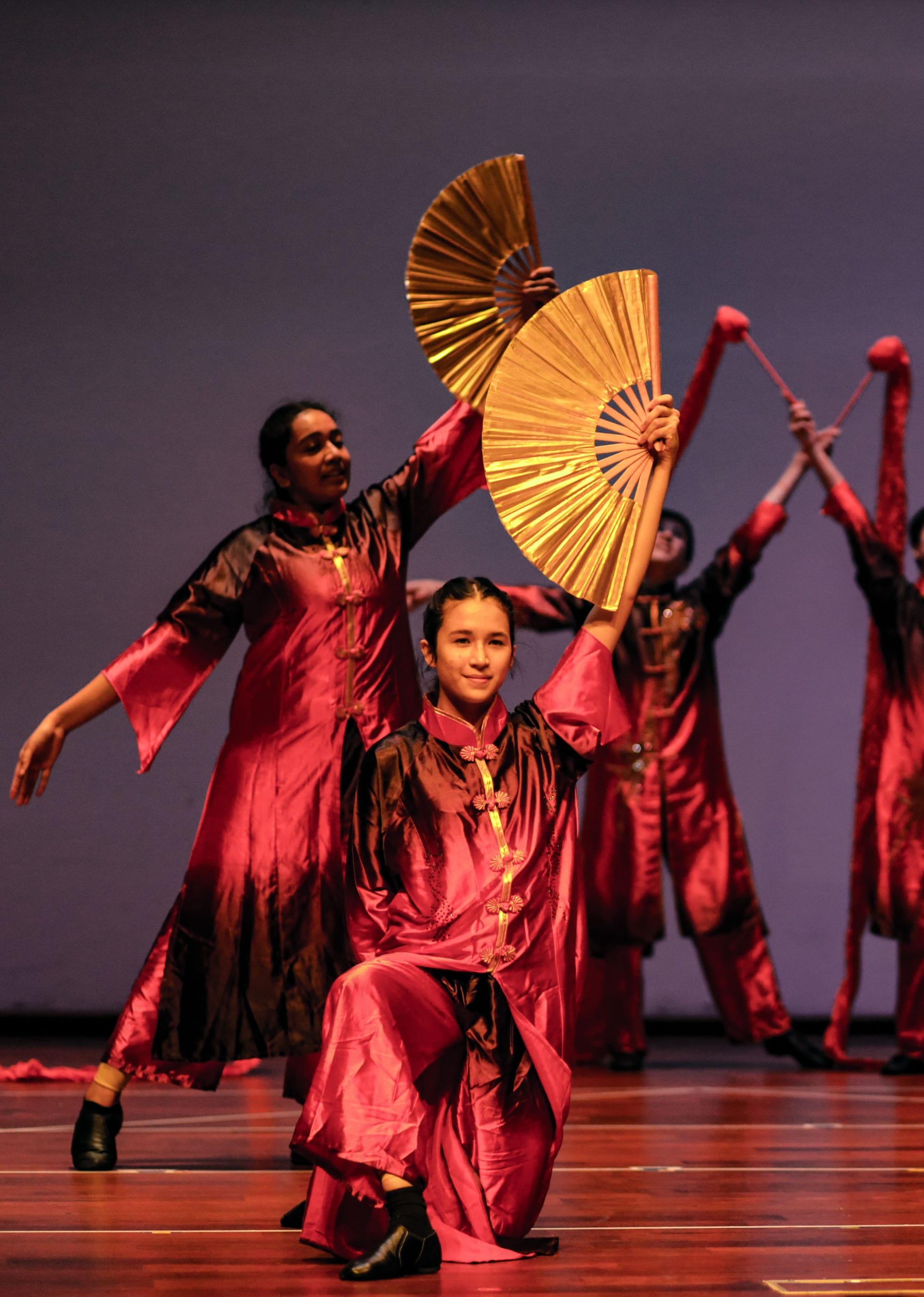 ACS(I) dance members perform a Chinese fan dance in red and gold costumes, with large golden fans fanned out dramatically on stage.