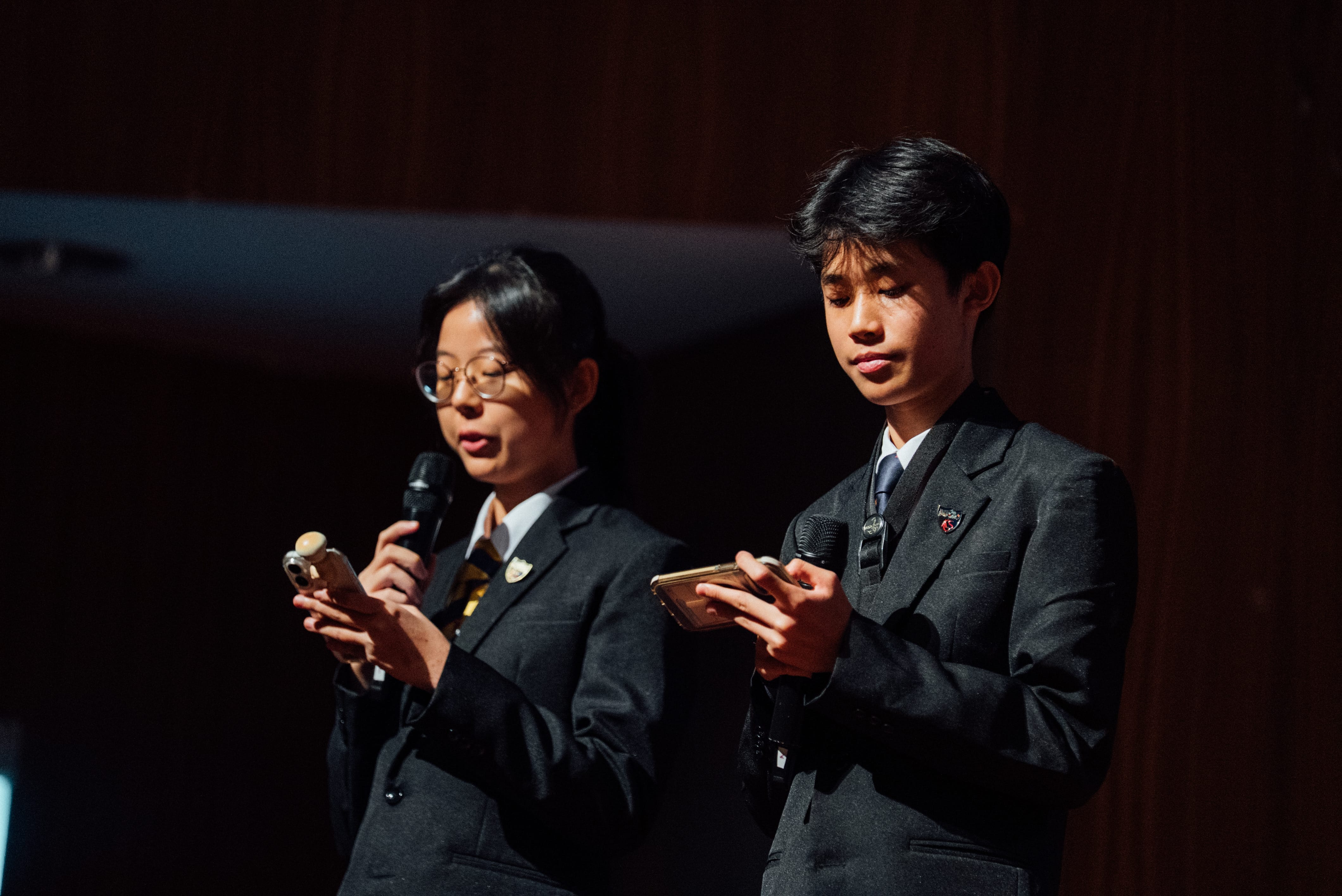 Two students in black blazers hold microphones and look at their phones on a dark stage.