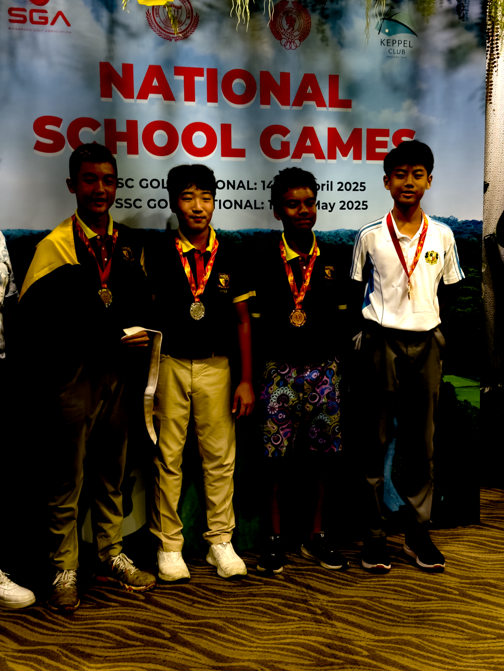 Four young male students wearing medals stand before a National School Games golf banner at Keppel Club, 2025.