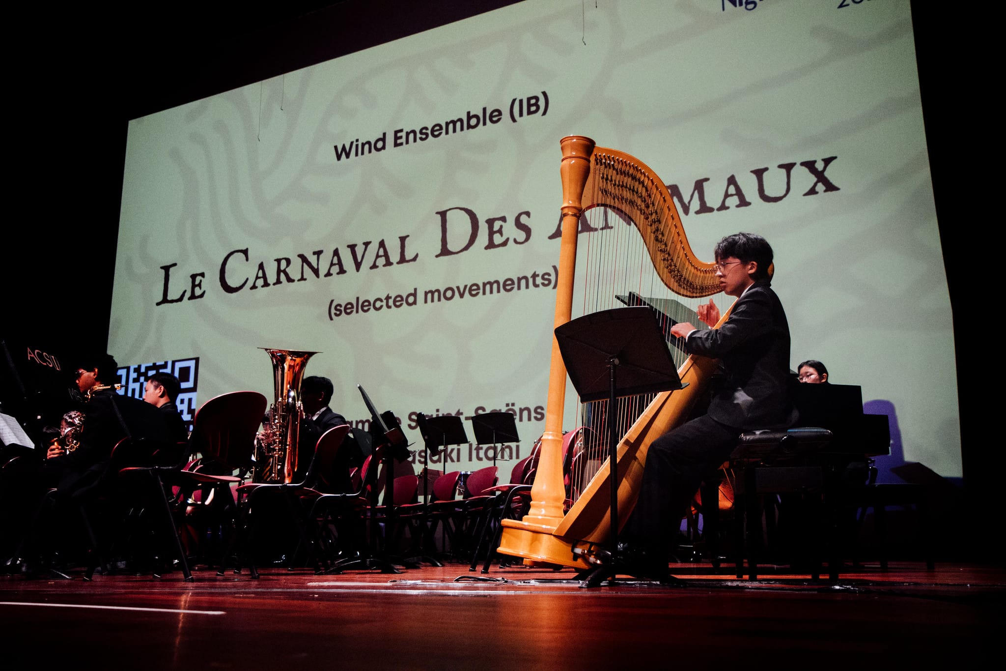 A harpist performs on stage with a wind ensemble, a screen behind reads "Le Carnaval Des Animaux, selected movements."