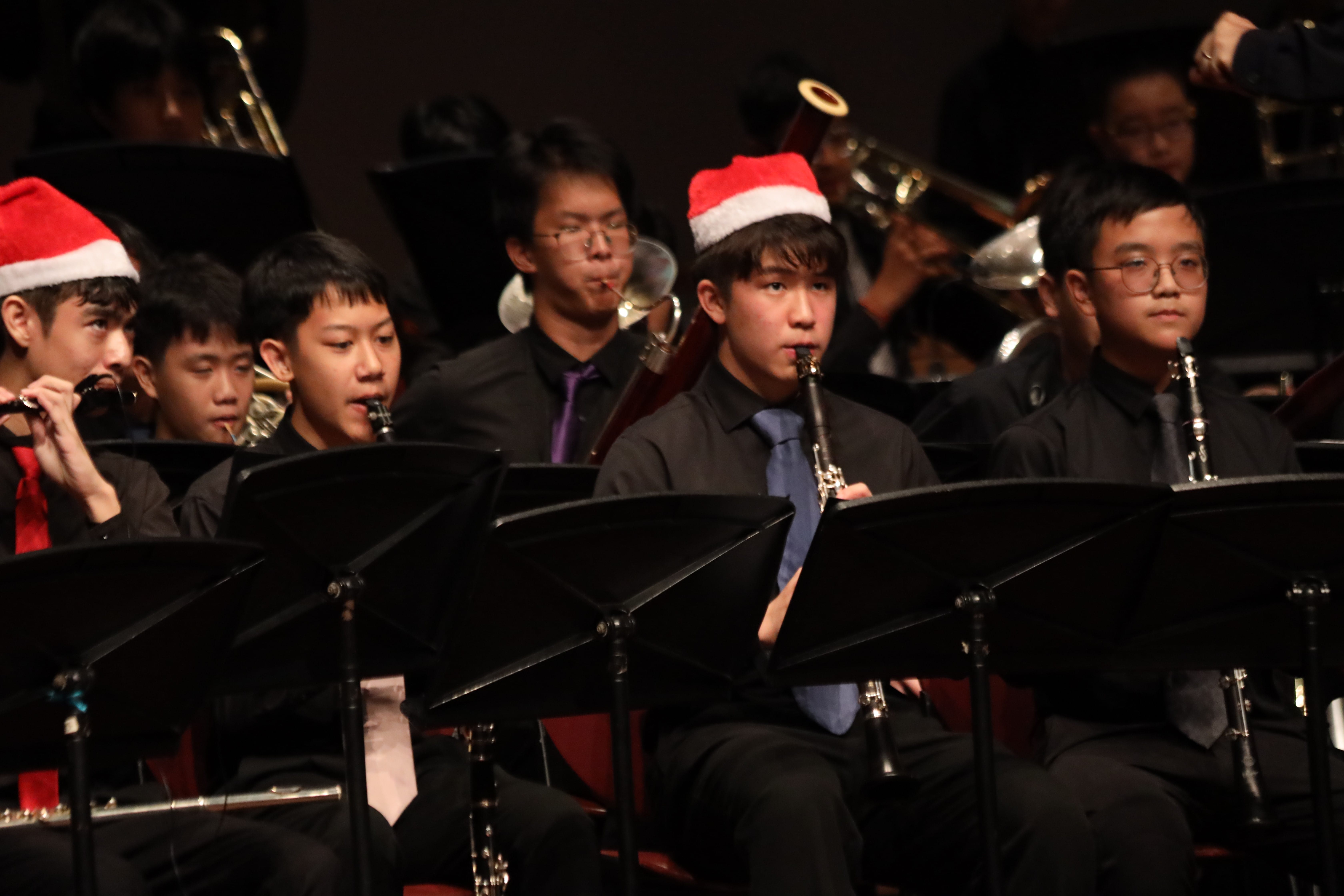 Young musicians in black attire and Santa hats play clarinets and brass instruments at a Christmas concert.