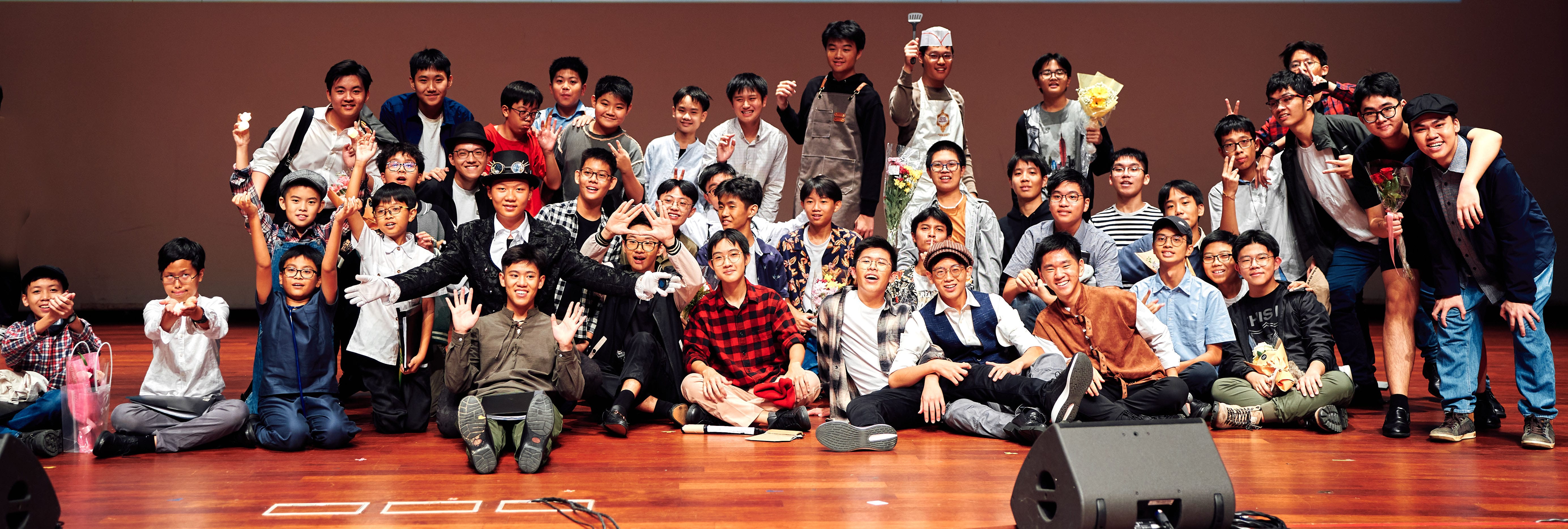 Large group of young Asian males posing cheerfully on a wooden stage, some holding flowers, waving and smiling.