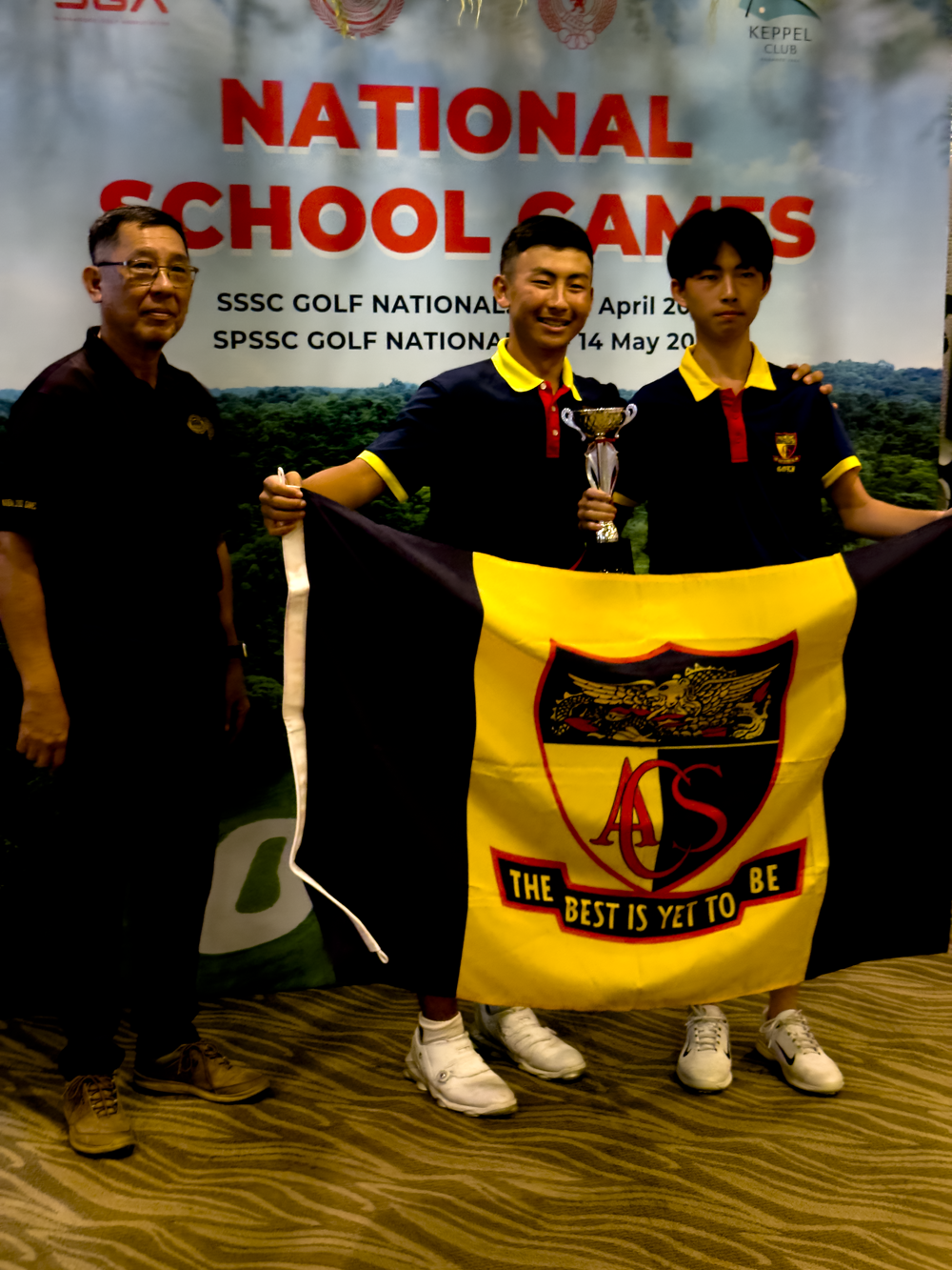 Three people pose with a trophy and ACS school flag at the National School Games SSSC Golf Nationals banner.