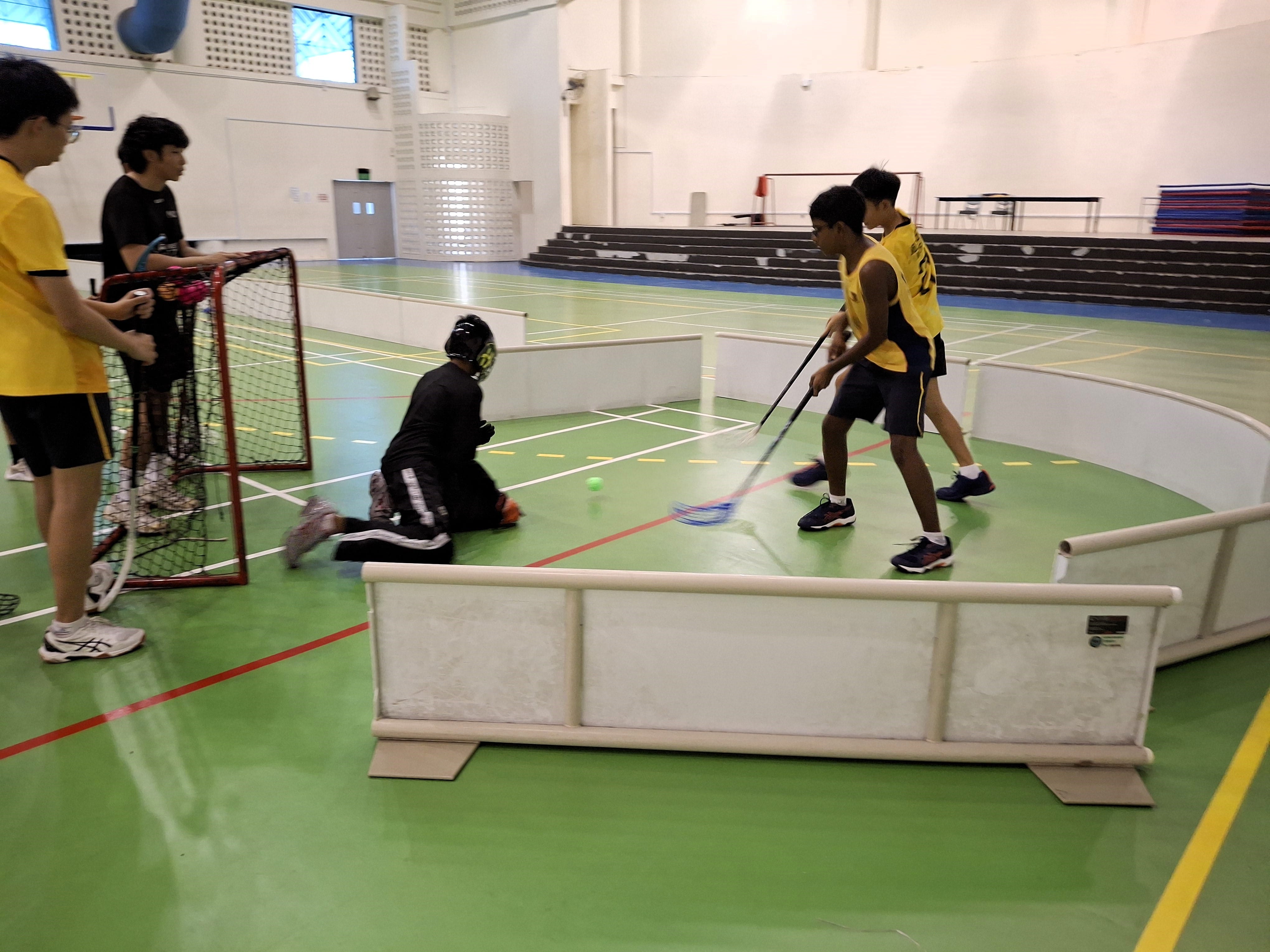 Boys playing floorball indoors, with two players in yellow shooting at a goal defended by a kneeling goalkeeper.