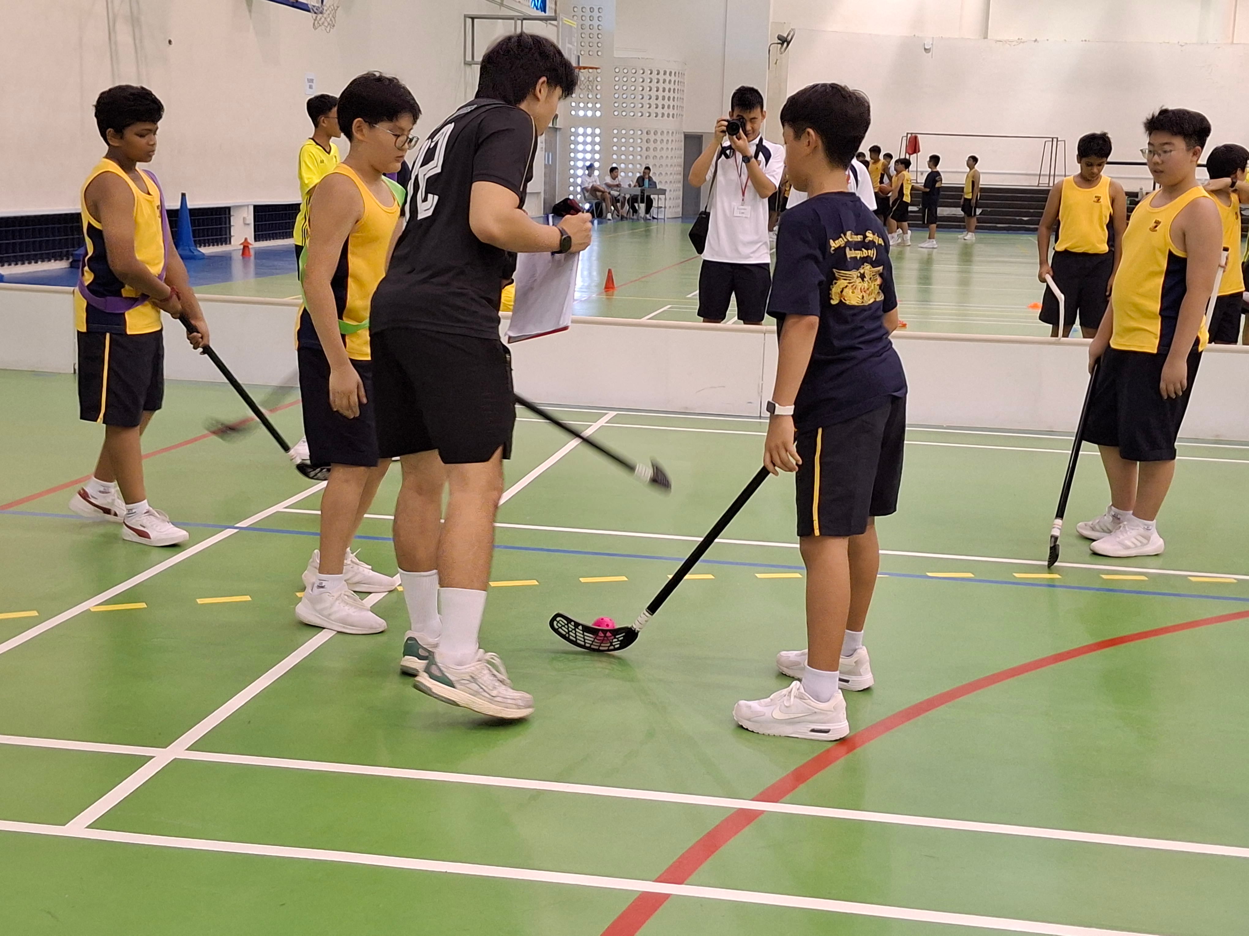 Students in yellow jerseys practice floorball with sticks and a pink ball inside a gym while a coach instructs them.
