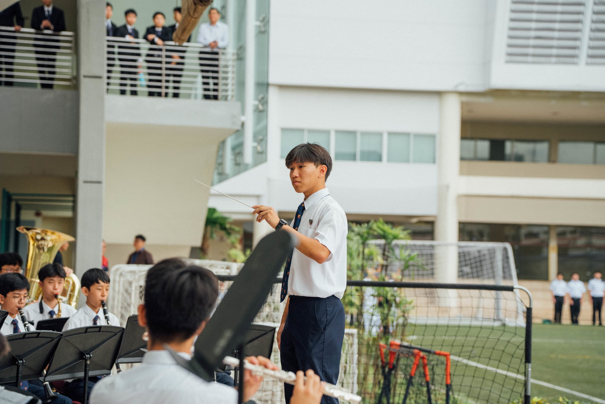 A uniformed student conducts a school band outdoors with a baton while others play instruments near a soccer goal.