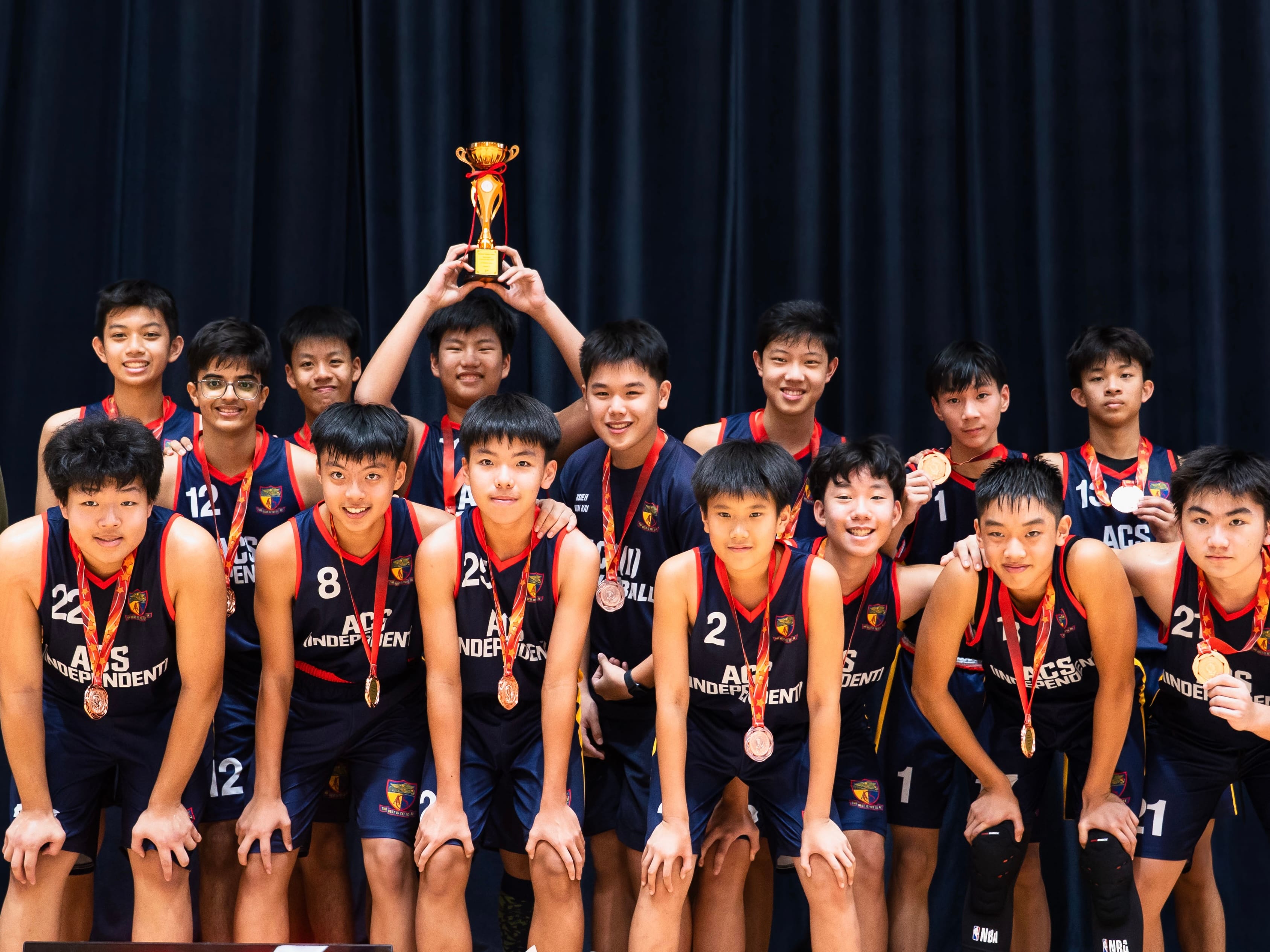 A youth basketball team in ACS Independent jerseys poses with medals and a gold trophy held overhead.