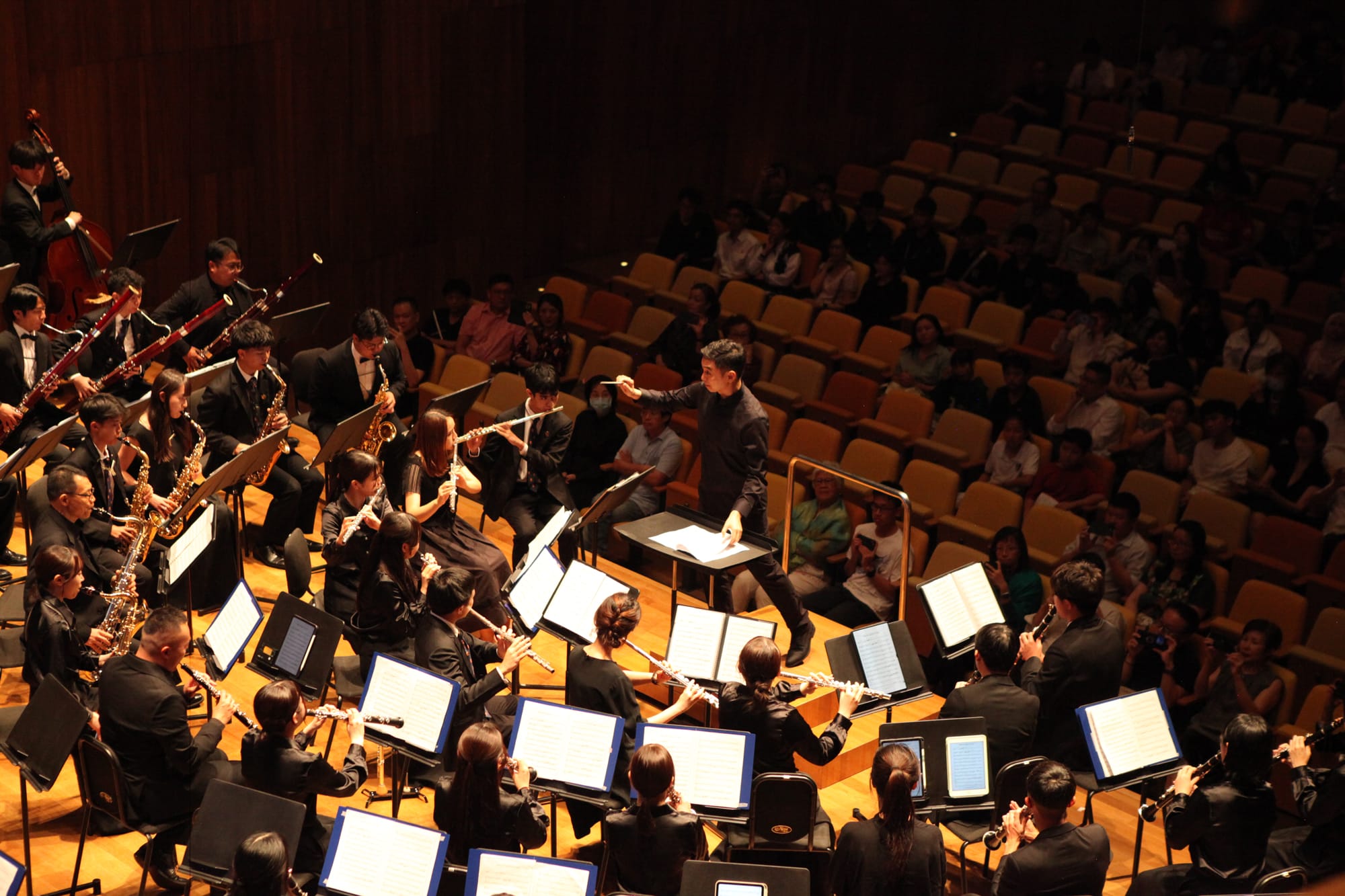 A conductor leads a large orchestra of wind and string players in black attire on a concert hall stage before an audience.