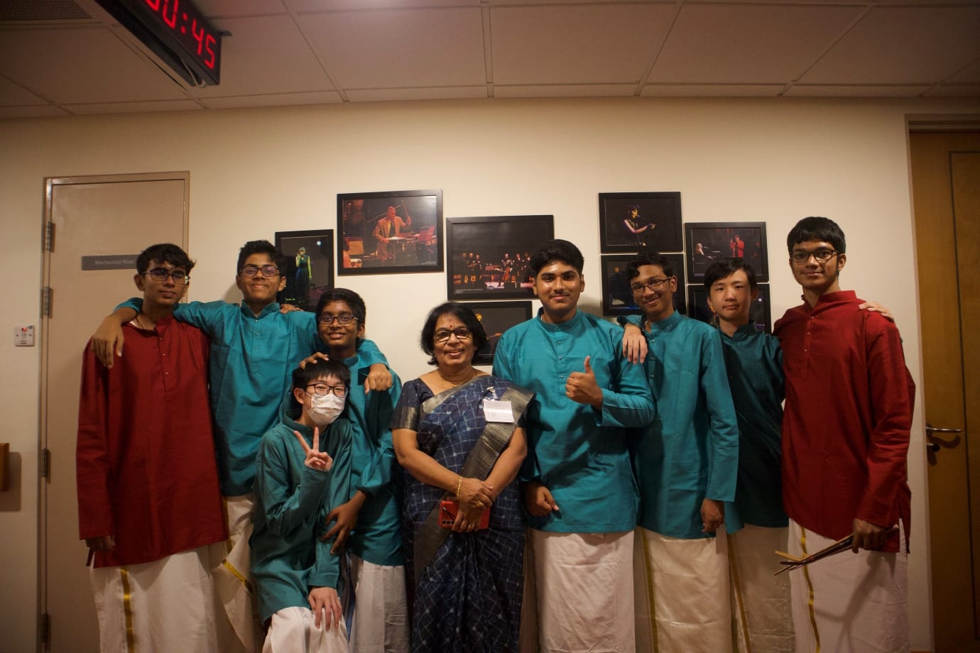 ACS(I) CPAC percussion members in teal kurta and white dhoti pose backstage with a teacher in a saree at a professional concert venue, smiling after a performance. Some members hold percussion sticks.