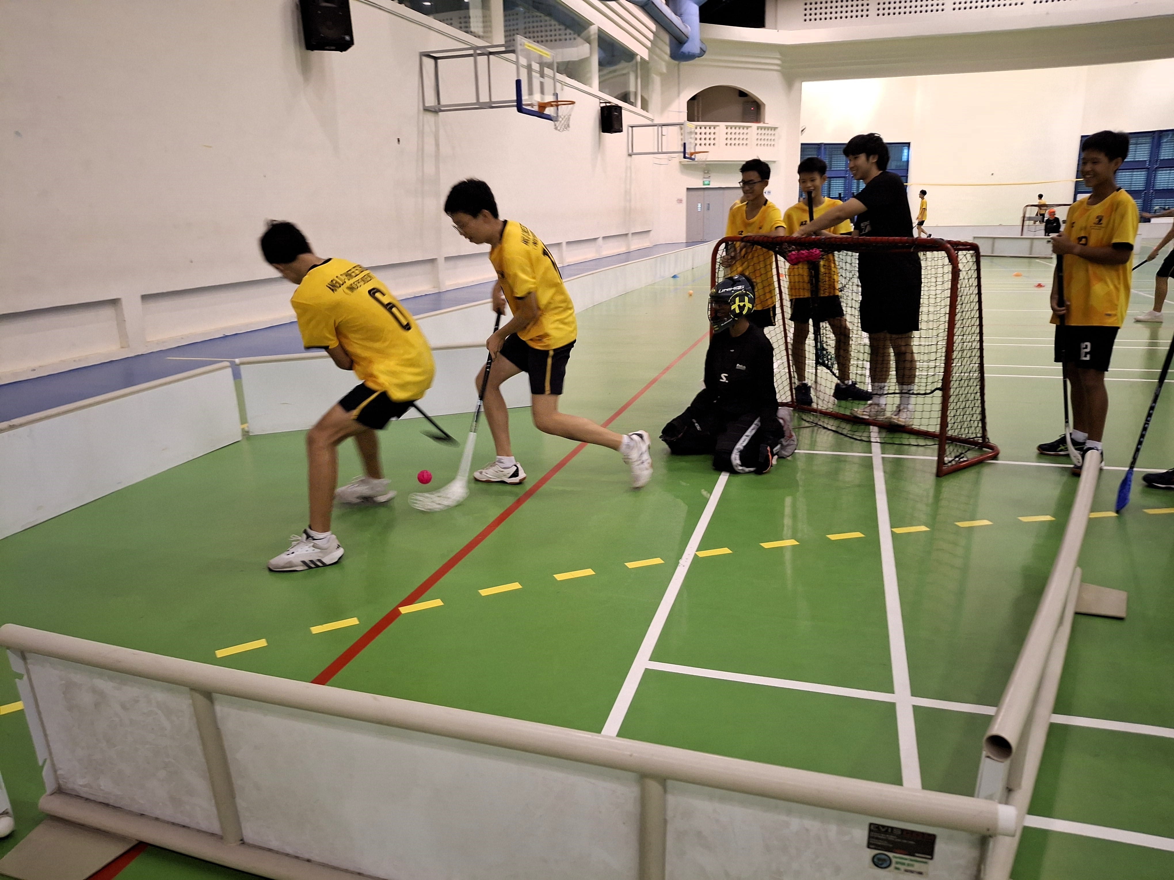 Youth players in yellow jerseys play indoor floorball on a green court, with a goalie in black gear defending a red net.