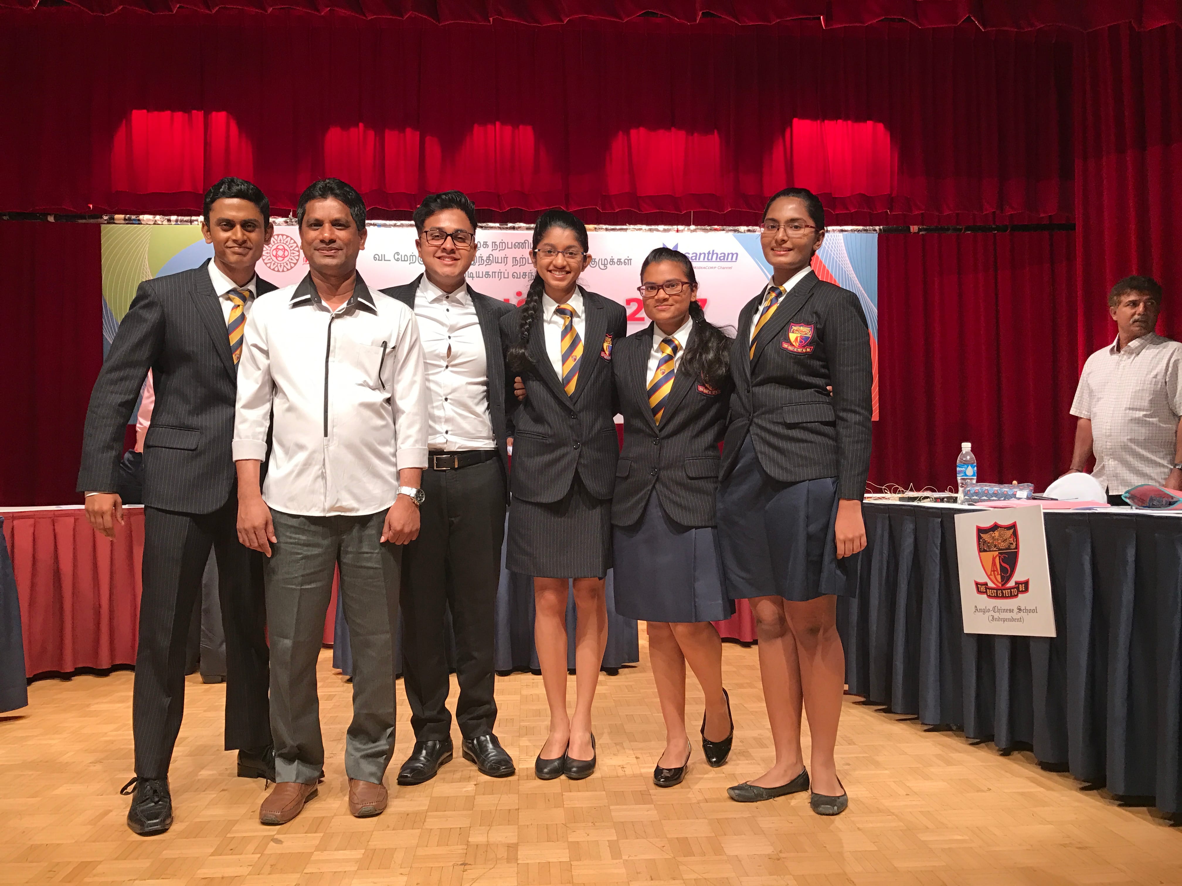 Seven people pose on a wooden stage before red curtains, three in school uniforms with striped ties and blazers.