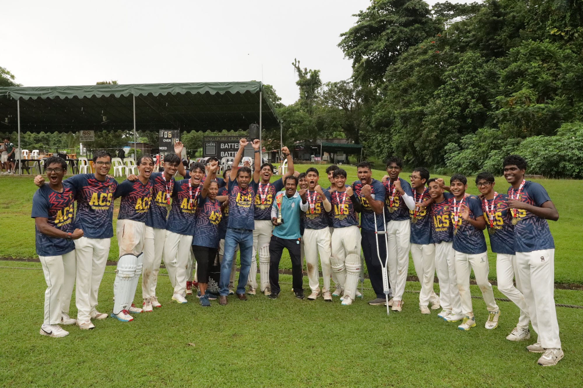 ACS cricket team celebrating on a field, wearing matching jerseys and medals, holding a trophy, cheering outdoors.