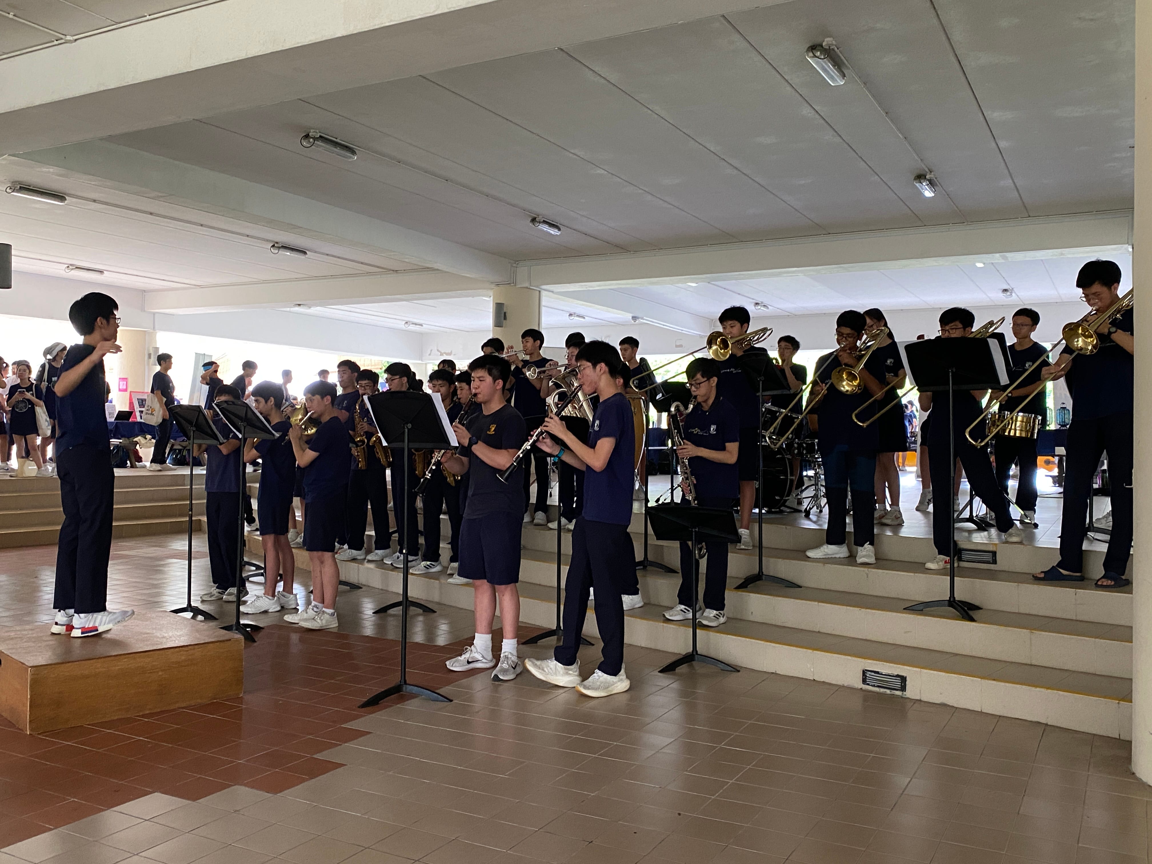 A youth brass band in navy uniforms performs indoors, led by a conductor standing on a wooden box.