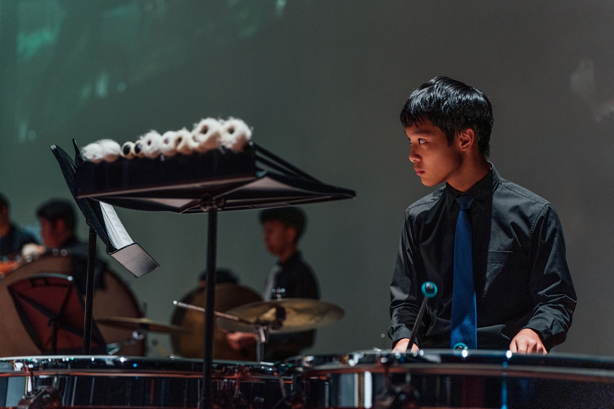 Young male musician in black shirt and blue tie playing timpani on stage with other performers in background.