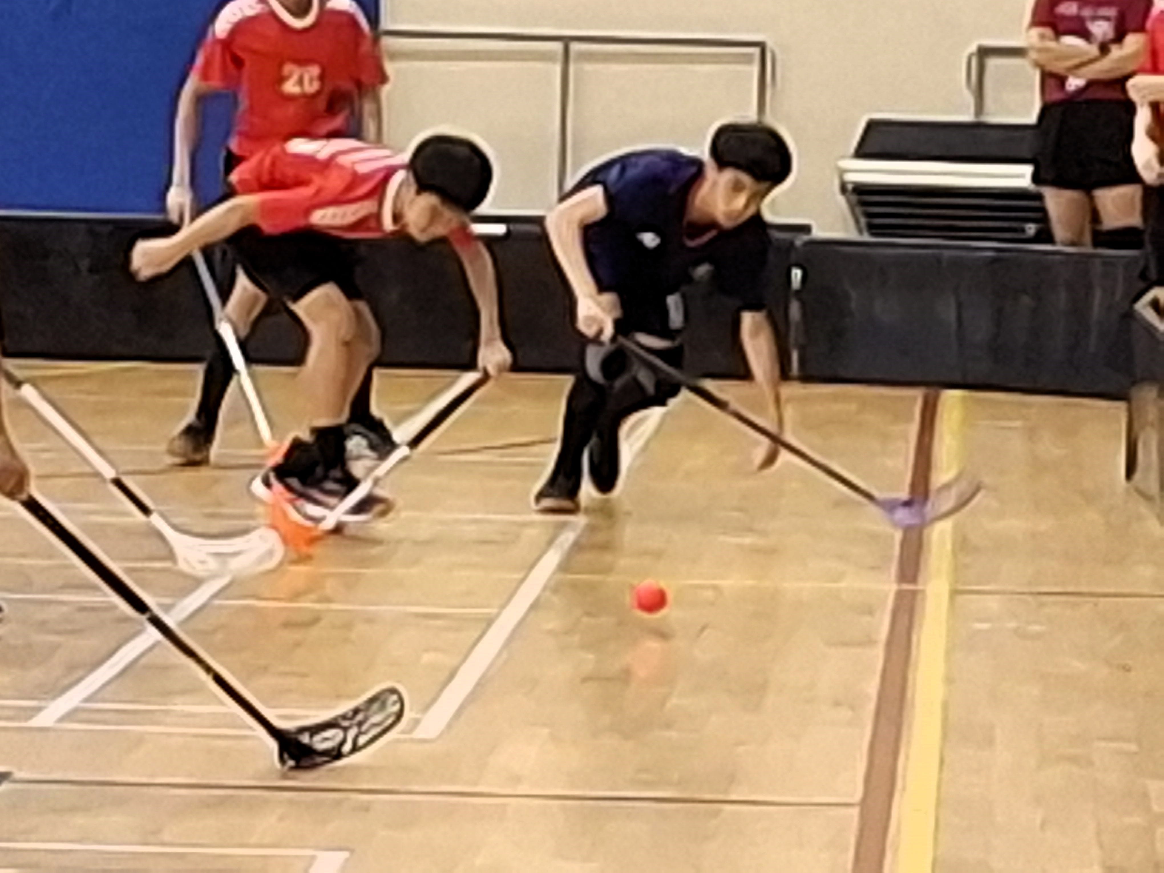 Two youth players compete for a small ball during an indoor floorball game on a gym floor.