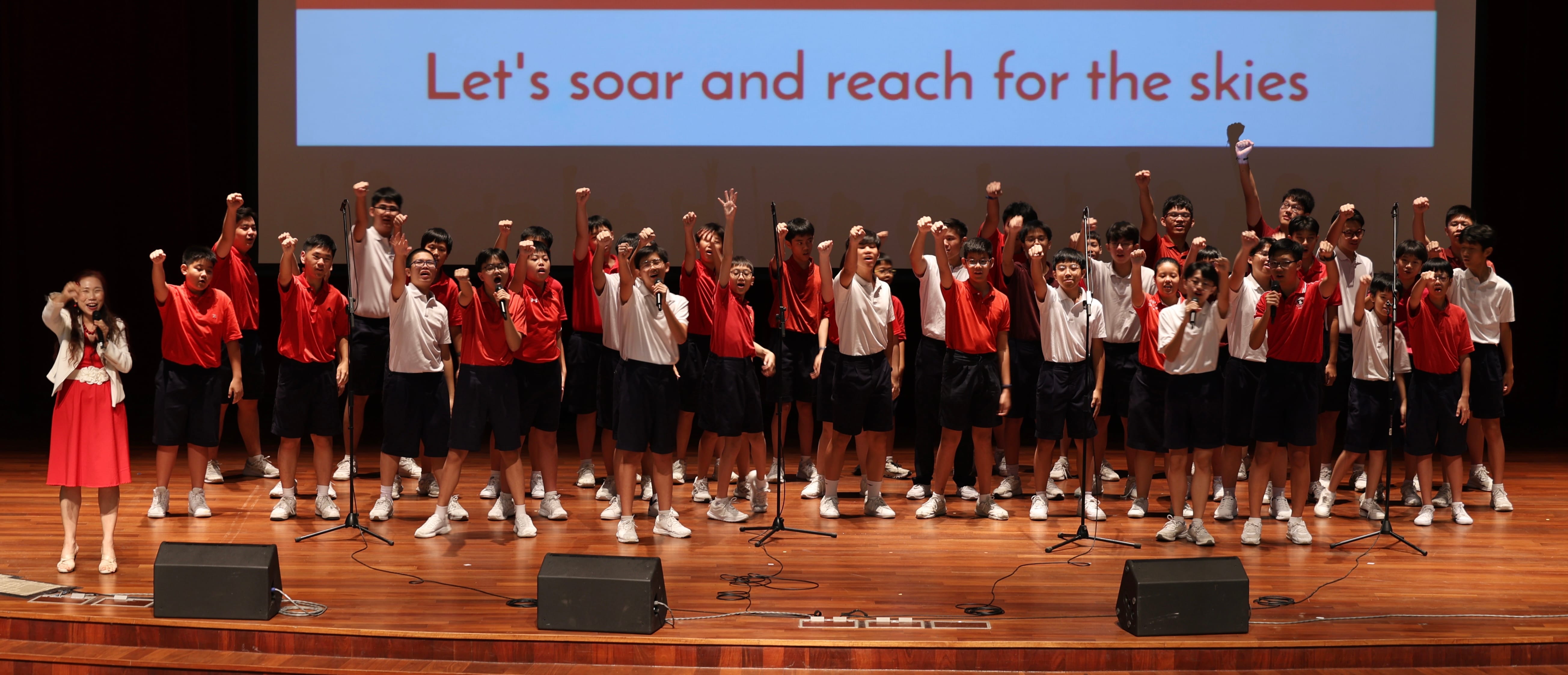 Students in red and white uniforms perform on stage with raised fists under a slide reading "Let's soar and reach for the skies."