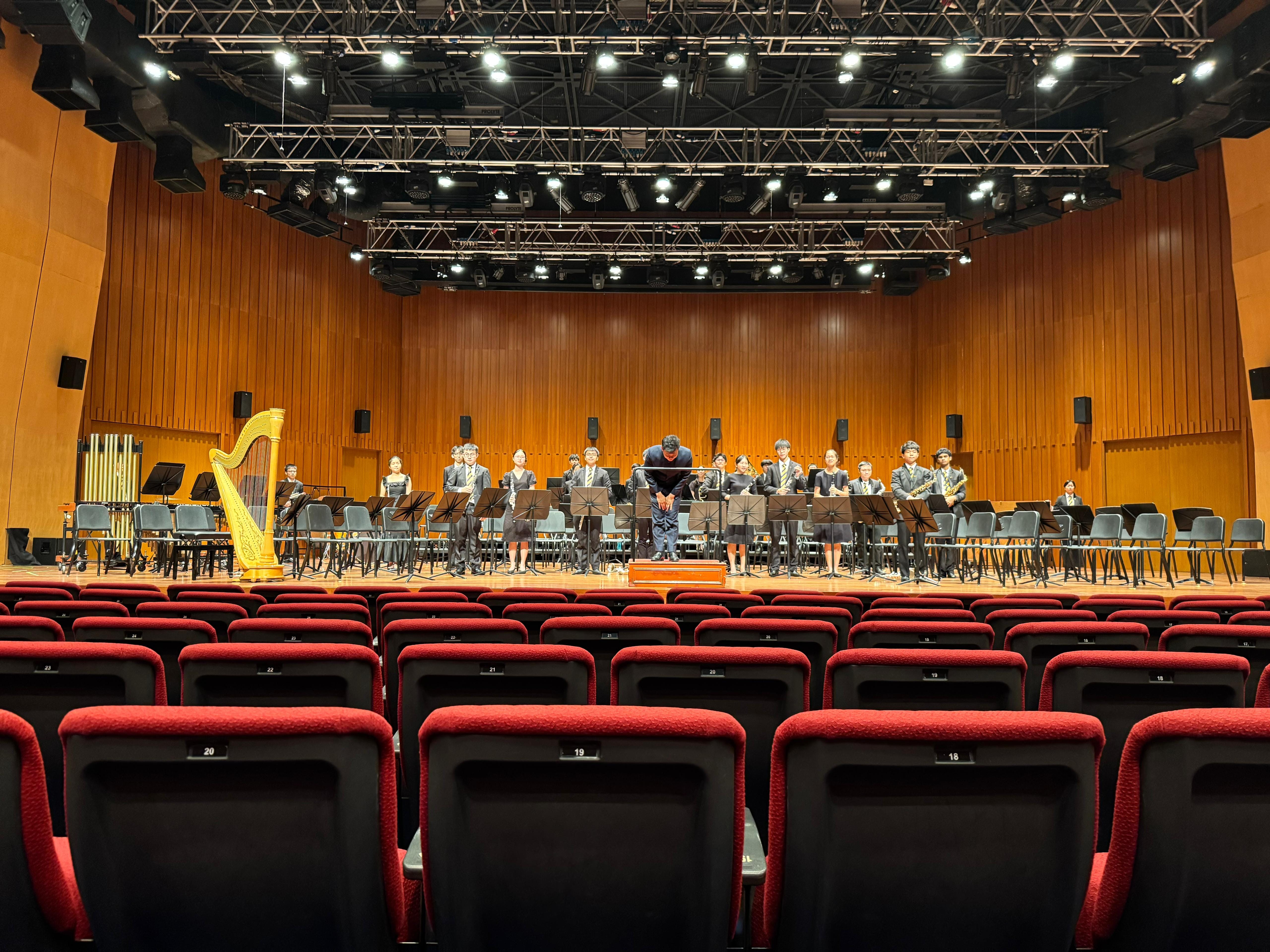 A youth orchestra stands on a wooden stage with a harp, music stands, and a conductor bowing before empty red seats.