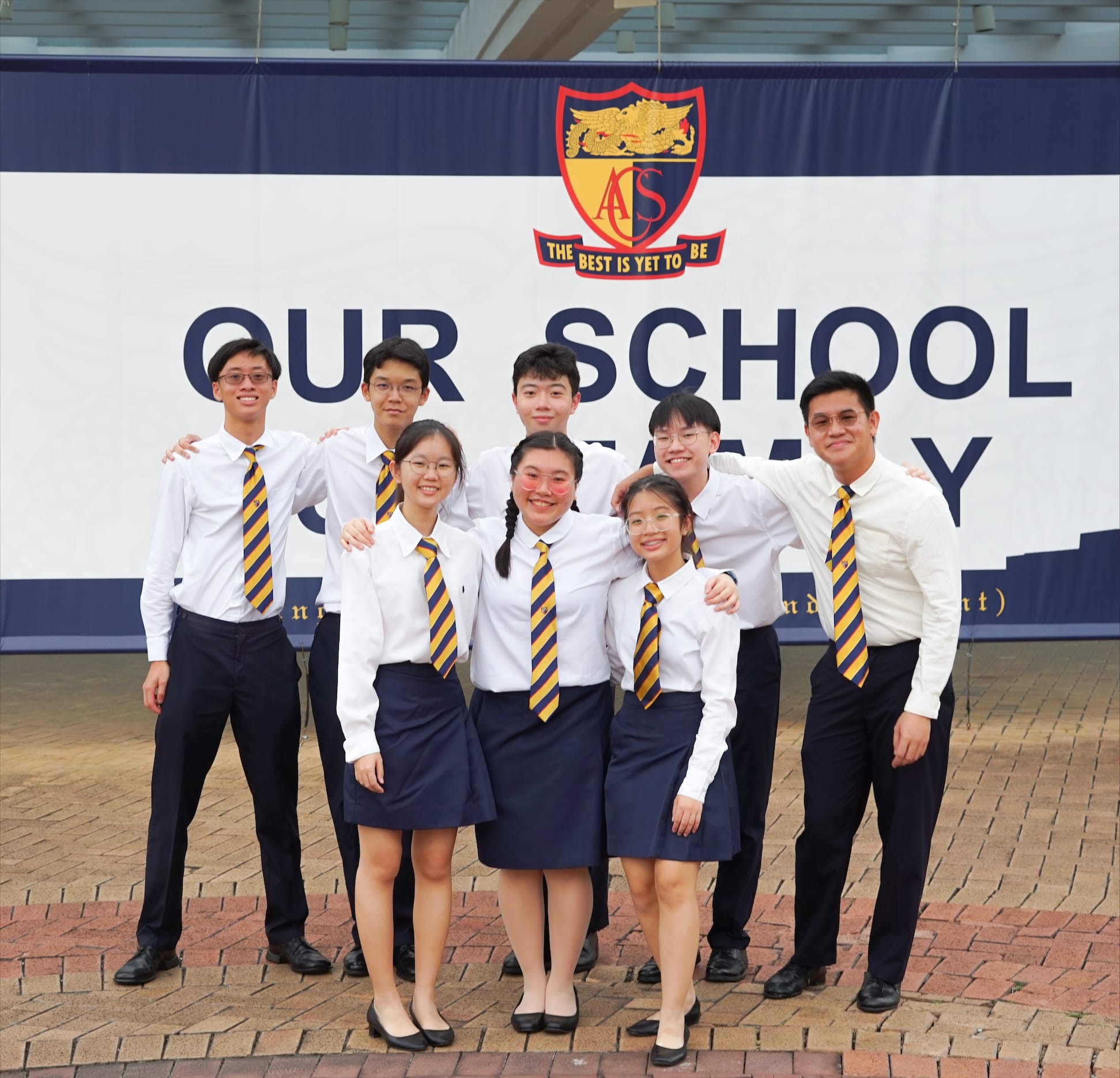Eight students in white shirts and navy striped ties pose smiling before an ACS school banner.