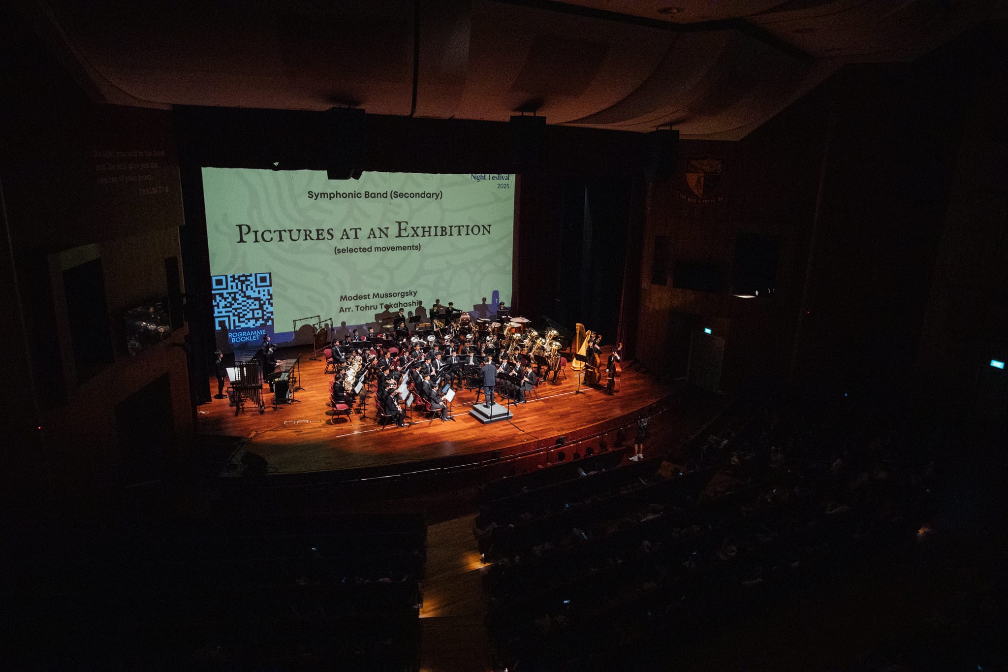 A symphonic band performs on a lit stage with a projected slide reading "Pictures at an Exhibition" in a dark concert hall.