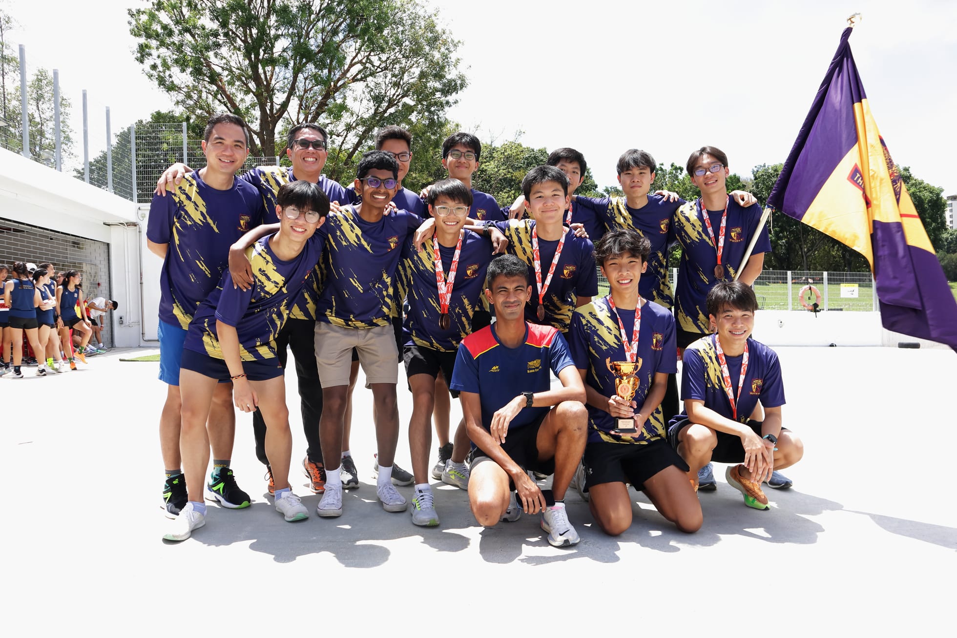 A youth sports team in purple and gold jerseys poses outdoors, holding a trophy and medals with a purple flag nearby.