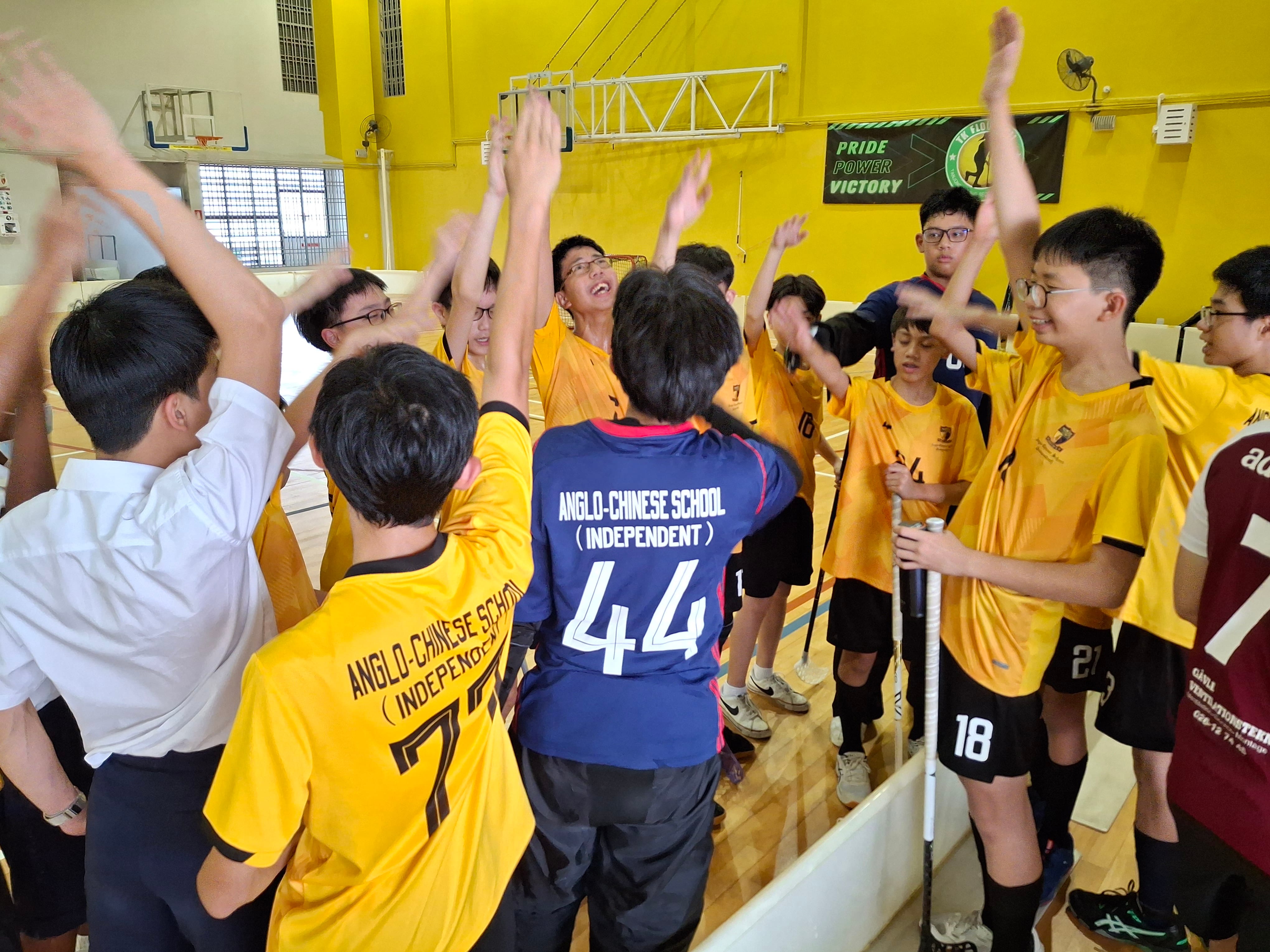 Anglo-Chinese School students in yellow jerseys raise hands in a team huddle inside a gymnasium.