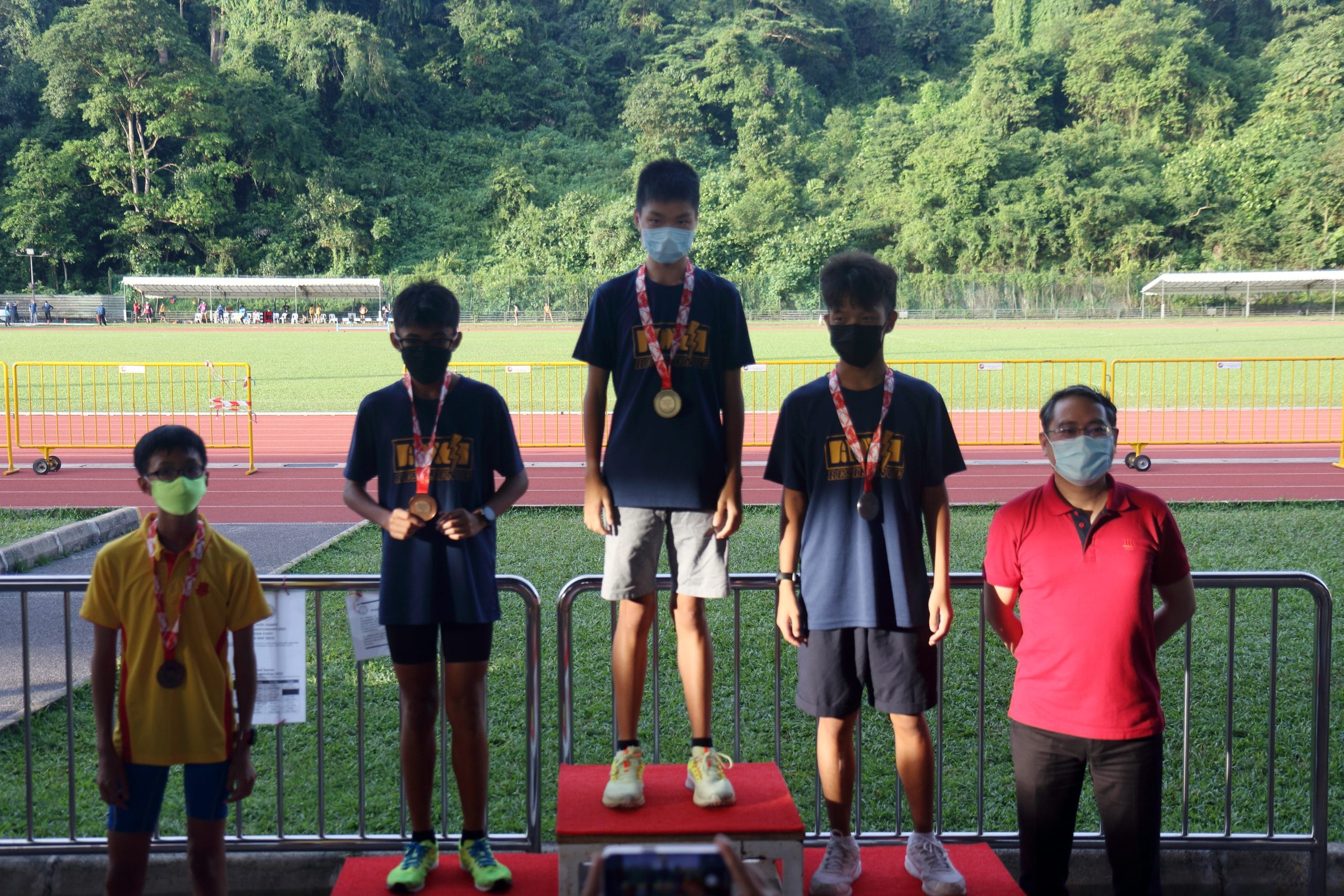 Four masked boys with medals stand near a podium at a track and field event, with an adult in red beside them.