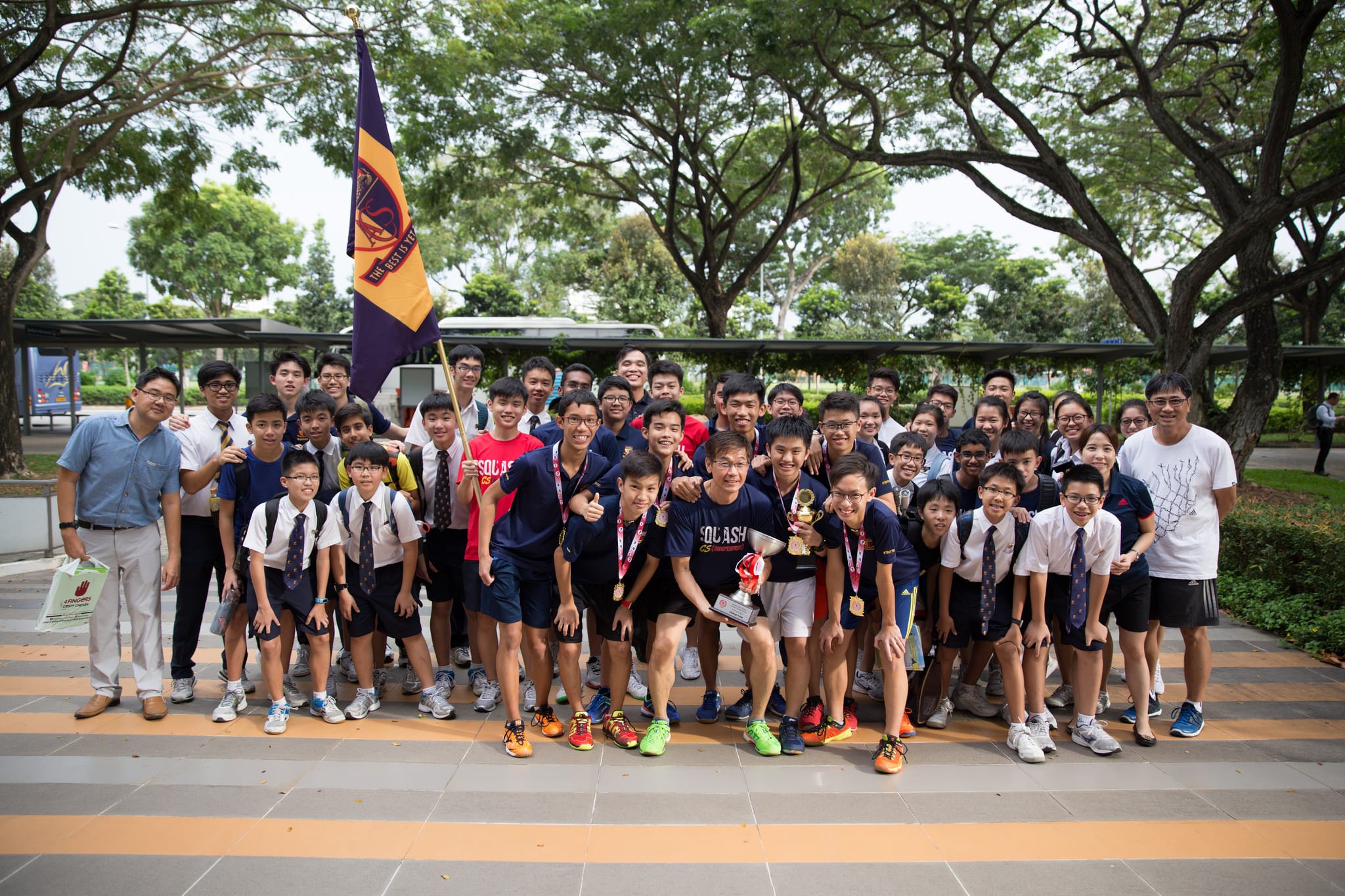 Large group of students and adults posing outdoors with a school flag and trophy, some wearing medals.