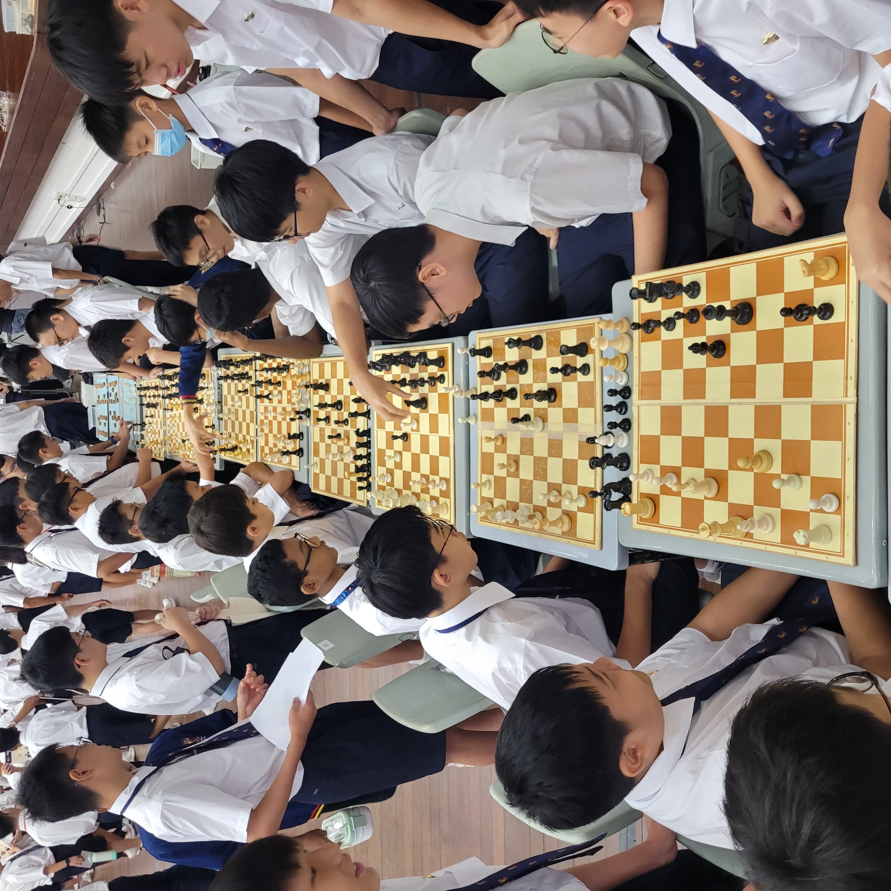 Students in white uniforms compete in a large chess tournament with multiple boards lined up in a row.