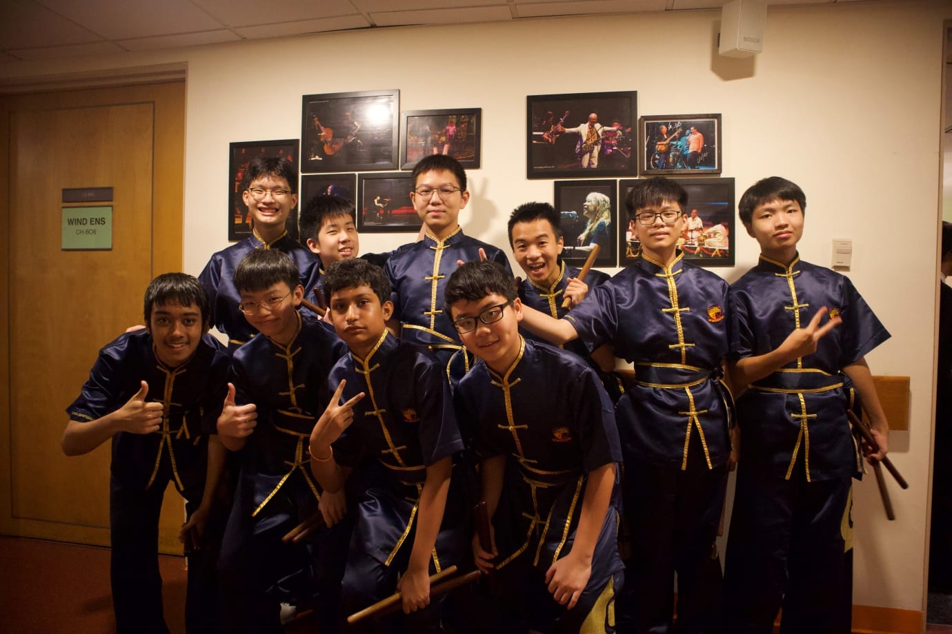 ACS(I) CPAC festival drum members in navy and gold uniforms pose cheerfully backstage outside a dressing room labelled "Wind Ens" at a professional concert venue, holding drumsticks and making playful gestures.