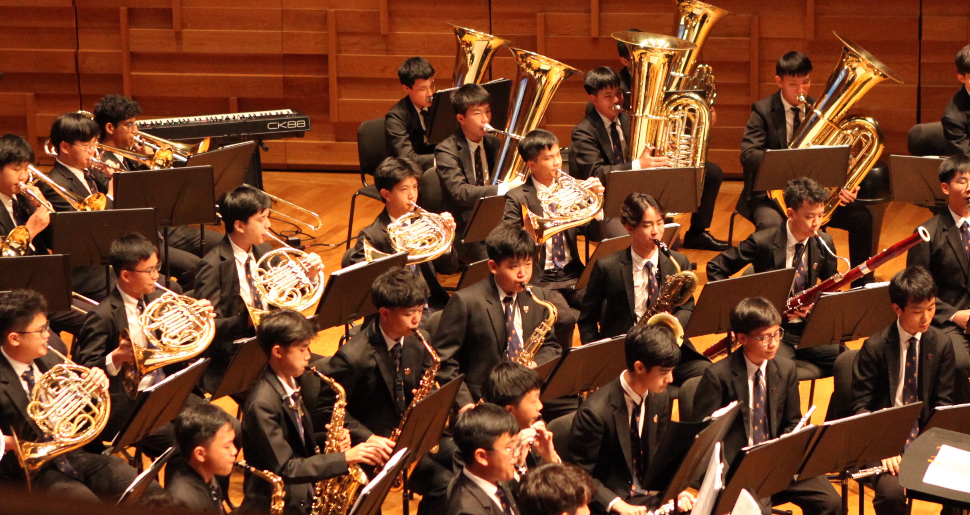 Young musicians in black suits perform in a large wind band, playing French horns, tubas, saxophones, and trombones on stage.