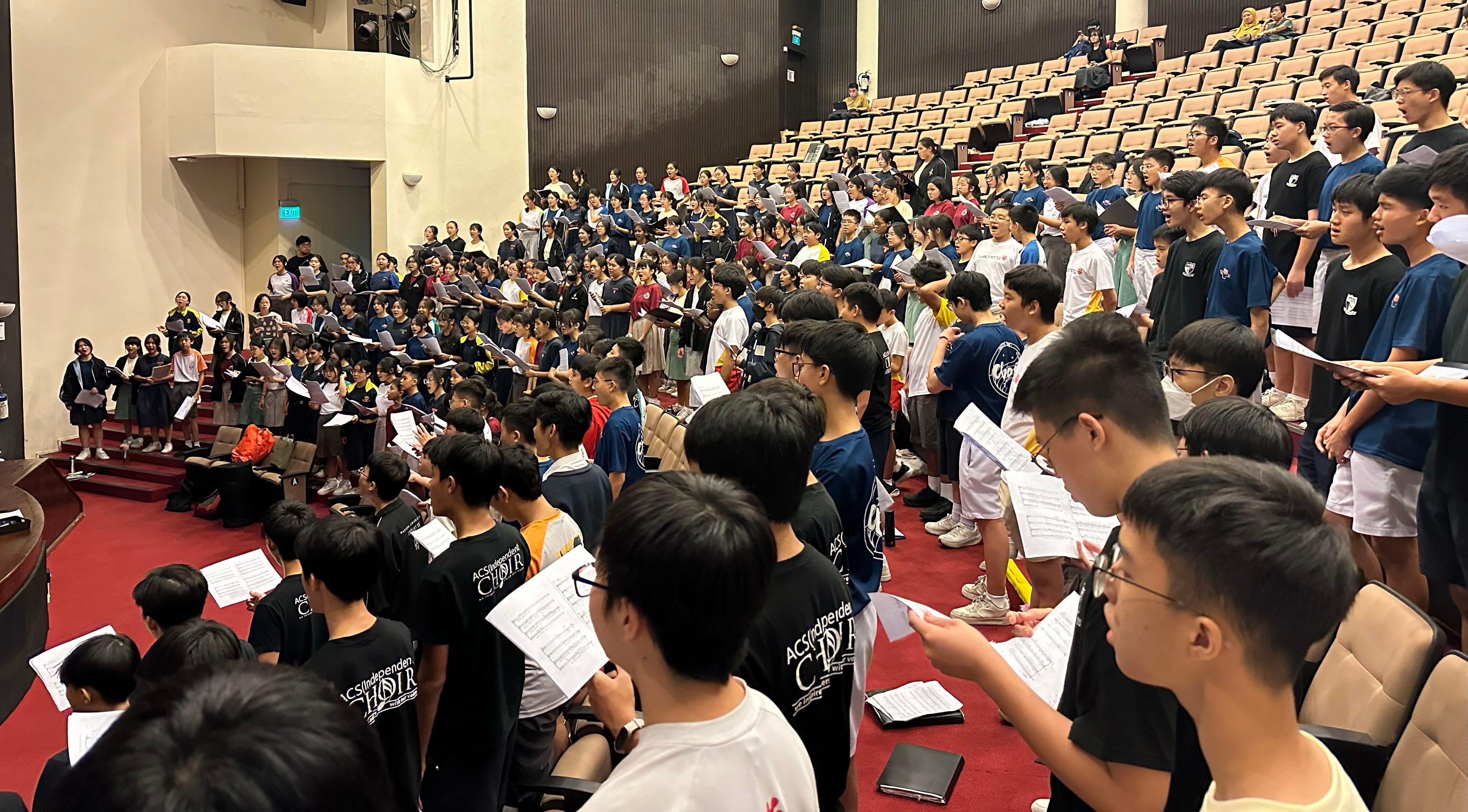 Large group of students in an auditorium holding sheet music, rehearsing as a choir on a red-carpeted stage area.