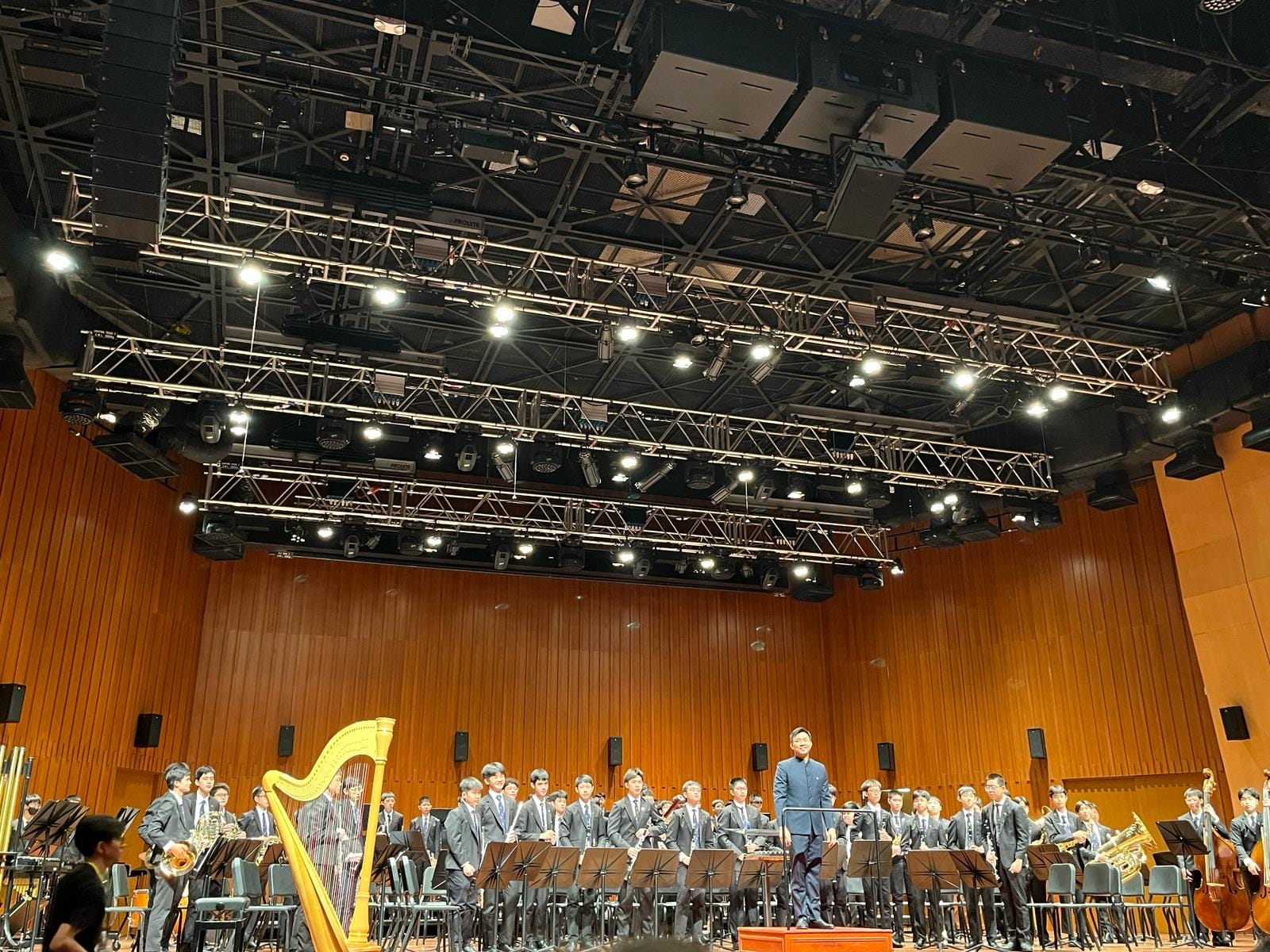 A youth orchestra in dark suits stands on a wood-paneled concert stage with a harp and conductor under bright stage lights.