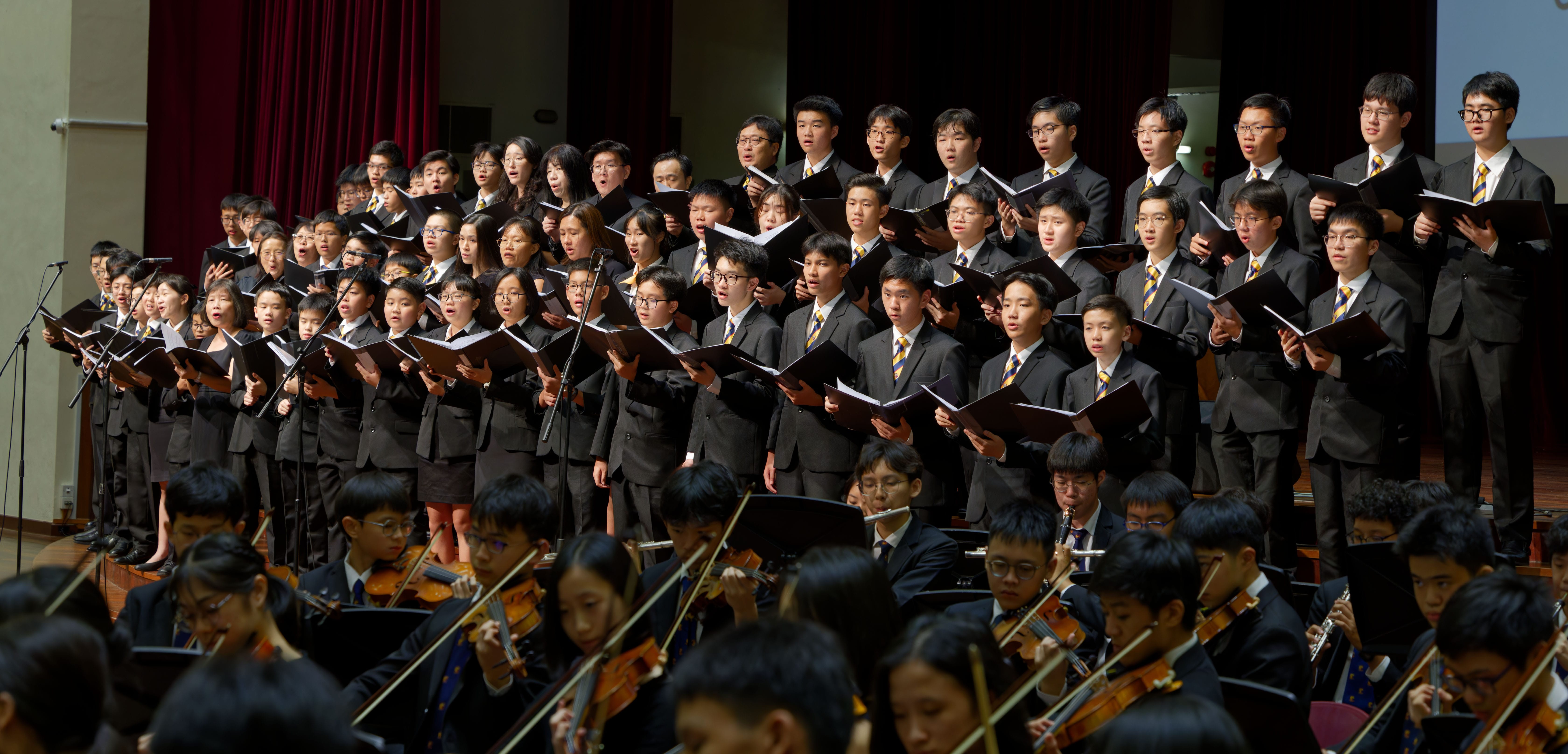 A large youth choir in dark suits holds music folders while an orchestra plays strings in front on a stage.