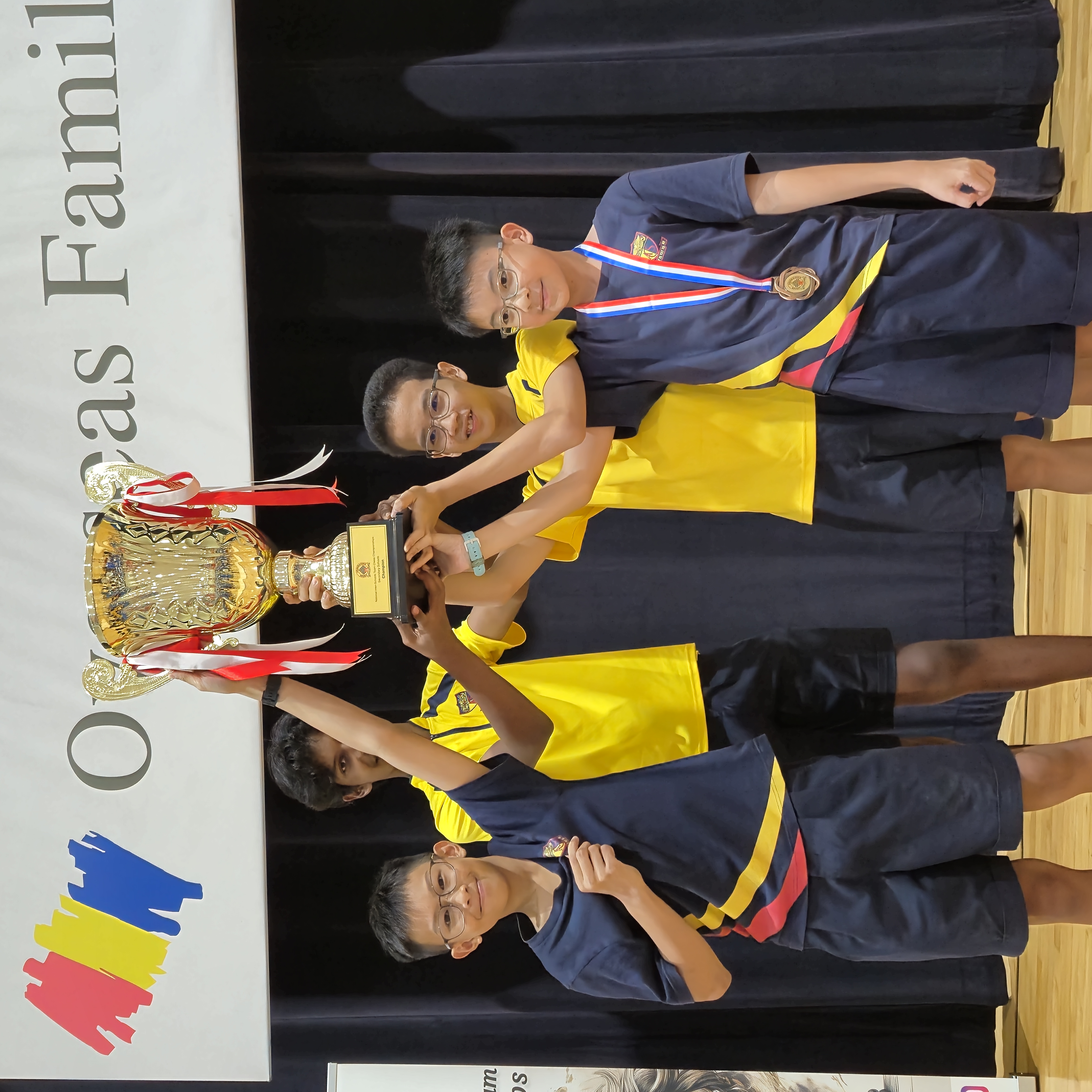 Four young boys in school uniforms lift a large gold trophy together on a stage under an Overseas Family banner.