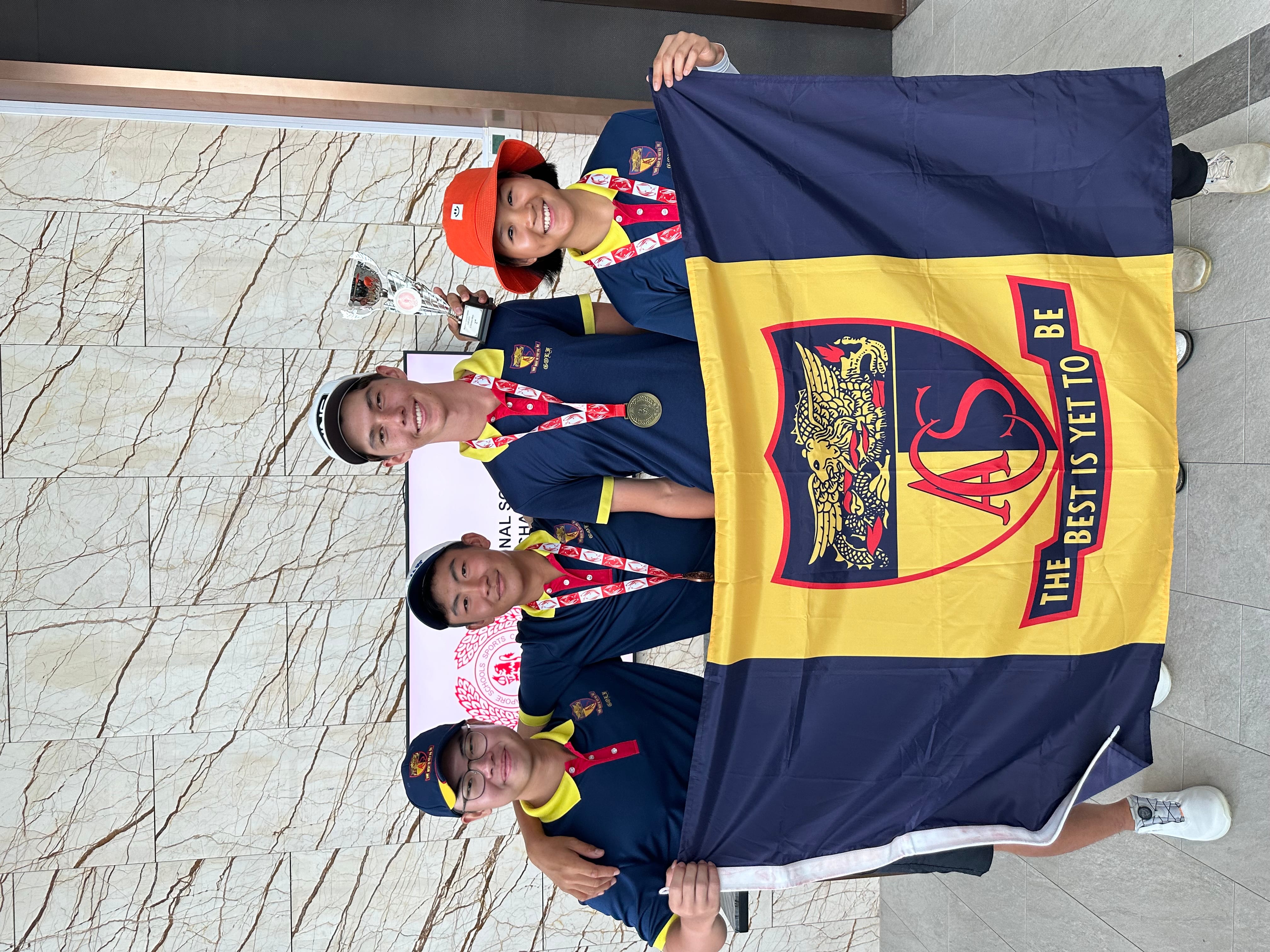 Four students in navy uniforms hold an ACS school flag and a trophy with medals around their necks.