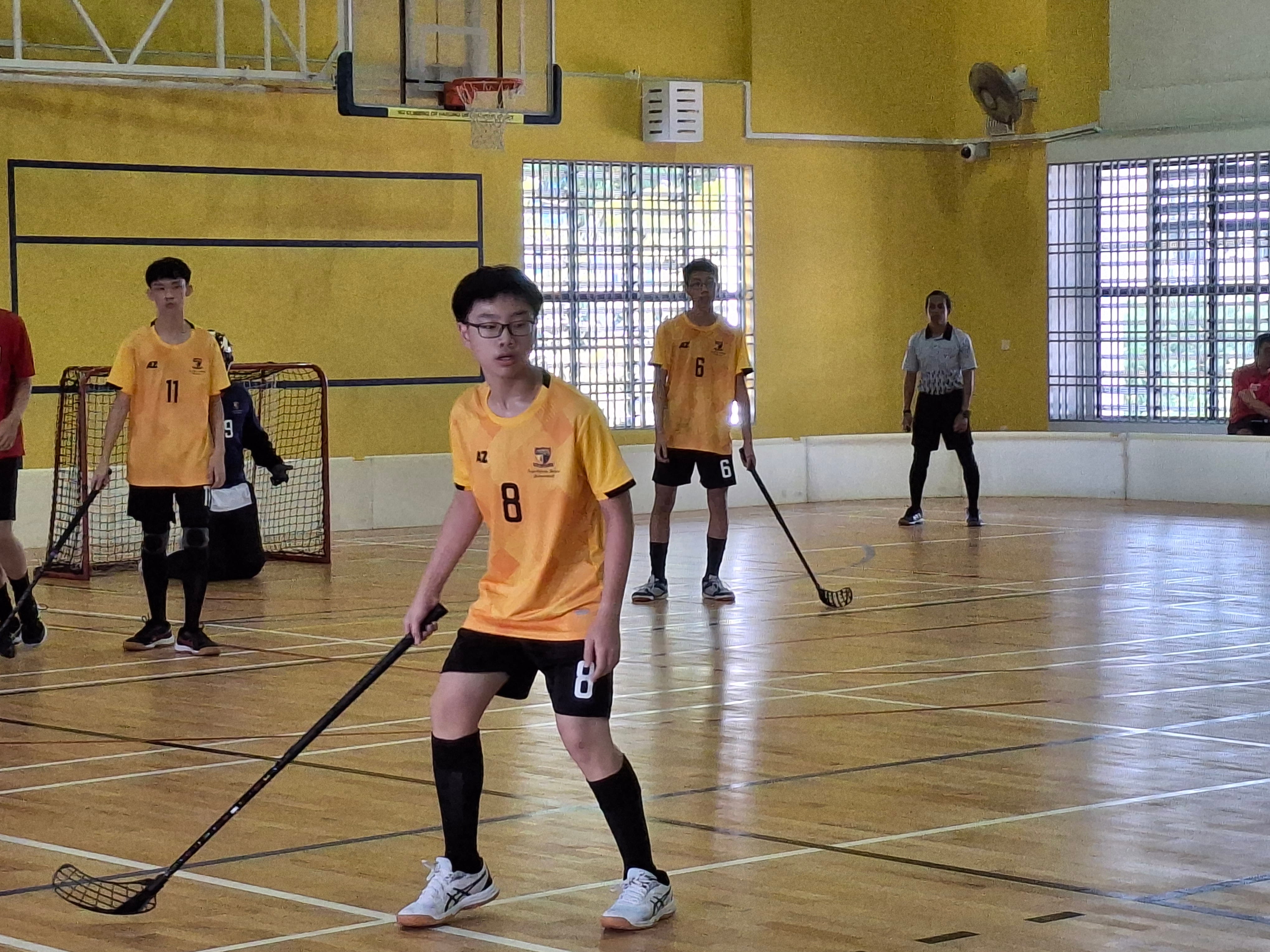 Young players in yellow jerseys holding floorball sticks on an indoor court with a goal net visible.
