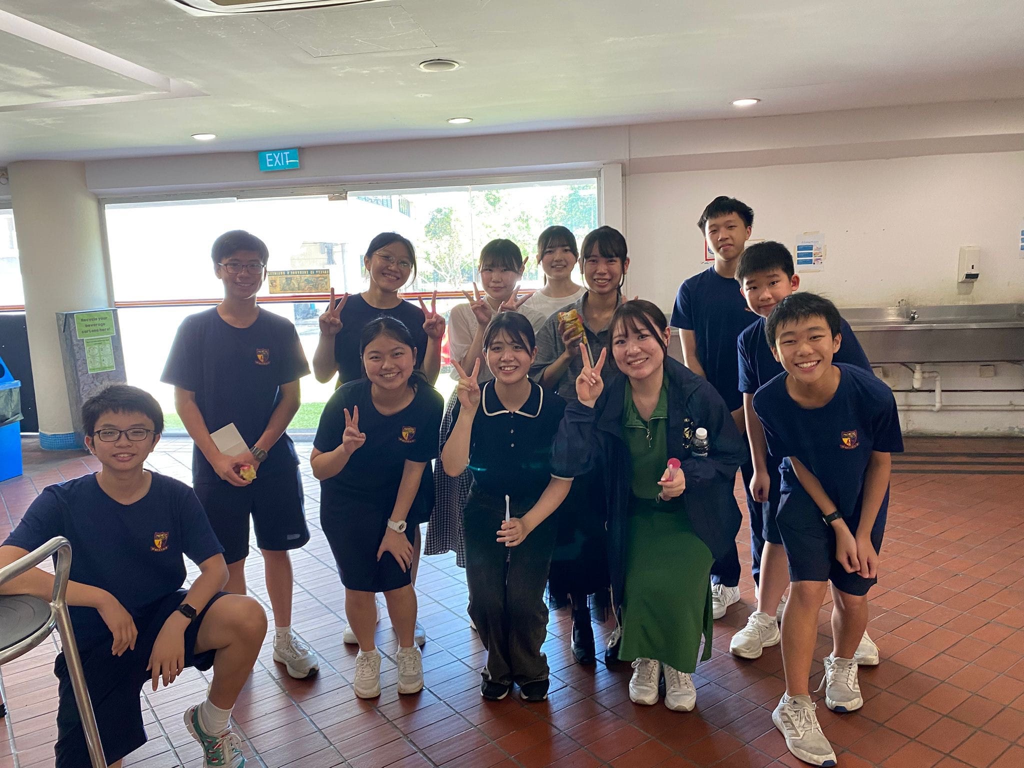 A group of smiling students in navy uniforms and two adults pose together indoors, several making peace signs.