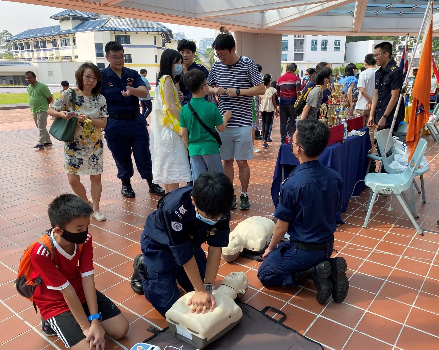 Two uniformed officers demonstrate CPR on mannequins outdoors while a crowd of adults and children watches.