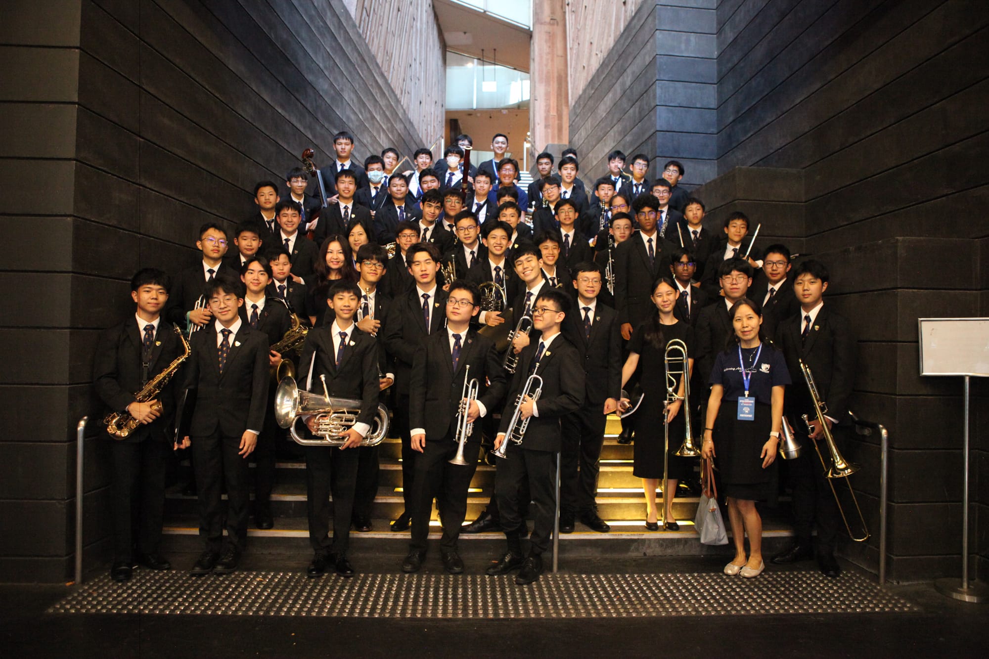 Large youth band in black formal attire poses on wide stairs holding brass and woodwind instruments.