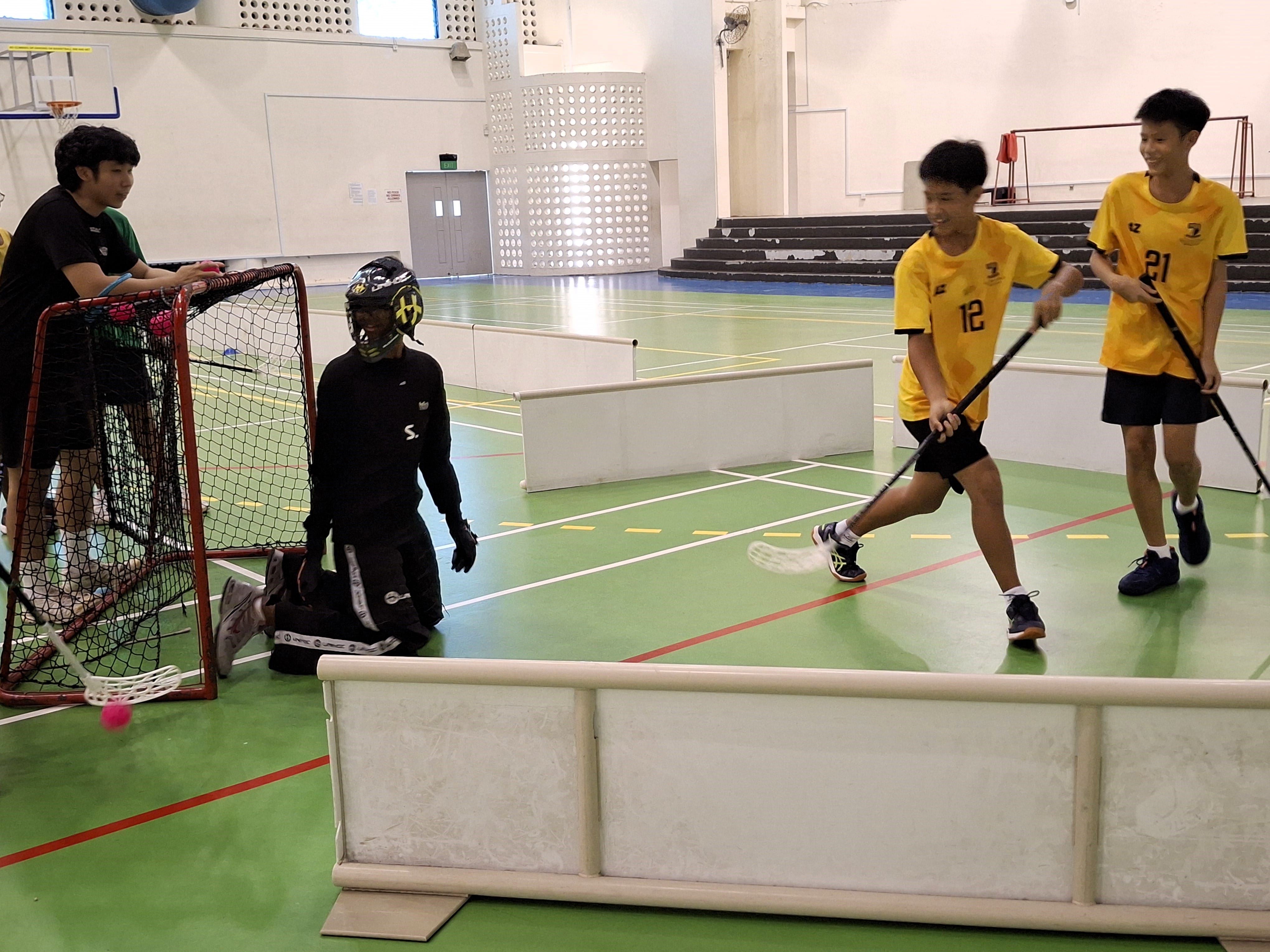 Young players in yellow jerseys play floorball indoors, with a helmeted goalkeeper guarding a red net.